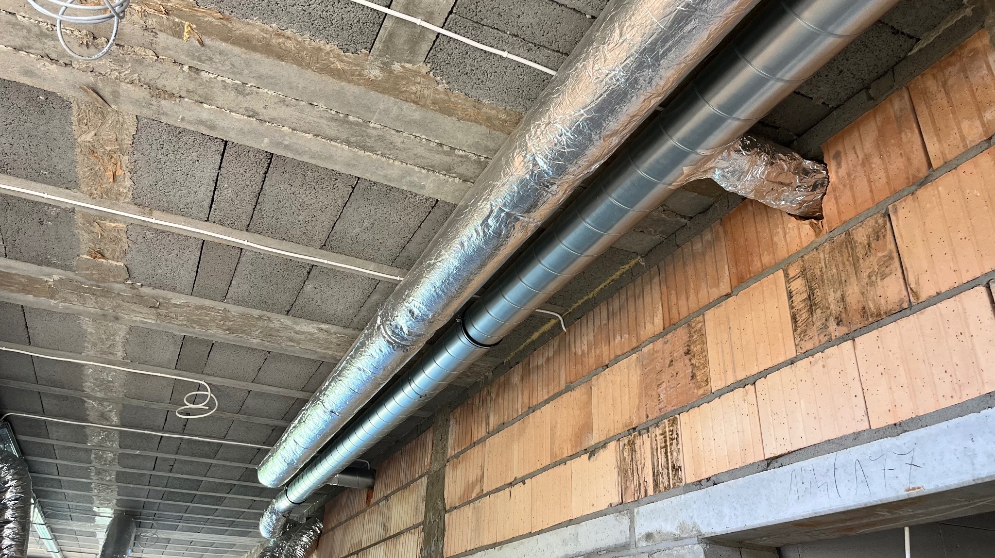 Surowe wnętrze z widocznymi przewodami wentylacyjnymi pod sufitem z betonowych bloków i ceglaną ścianą w tle. Widoczne łączenia i izolacje przewodów.