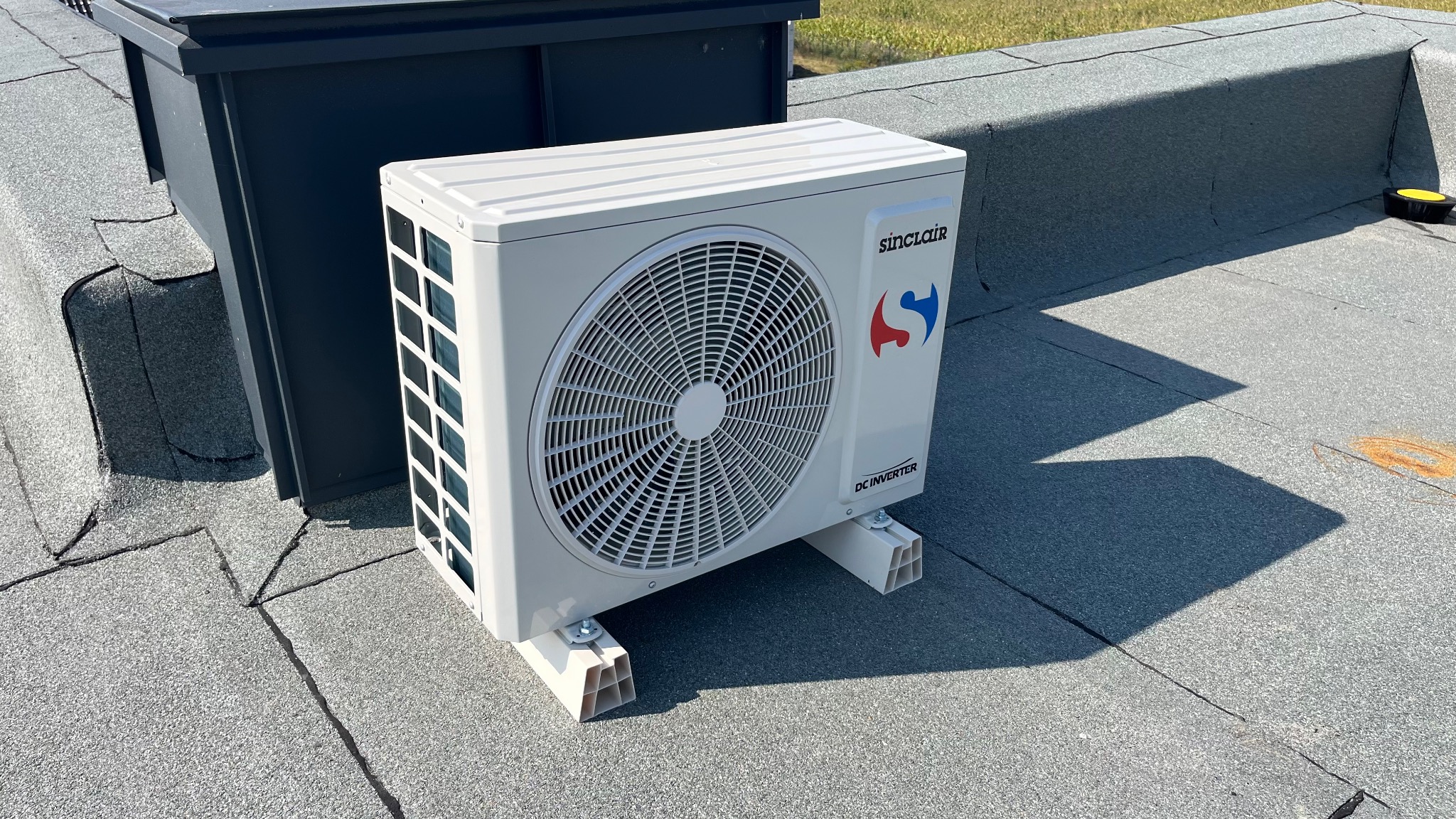 Biała jednostka zewnętrzna klimatyzacji Sinclair DC Inverter na płaskim dachu z szarej papy, obok ciemnej obudowy. Widoczne mocowania. Słoneczny dzień.