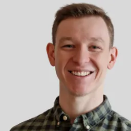 Uśmiechnięty mężczyzna w zielonej koszuli w kratę, portret z bliska na jasnoszarym tle. Widoczne detale twarzy i faktura materiału koszuli.