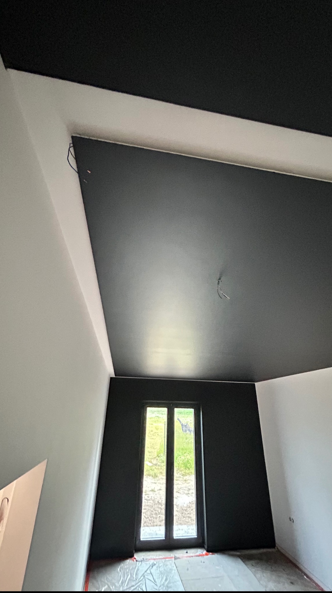 Wnętrze po malowaniu: czarny sufit i ściana z oknem, reszta ścian biała. Podłoga zabezpieczona folią malarską. Widoczne przewody na suficie.