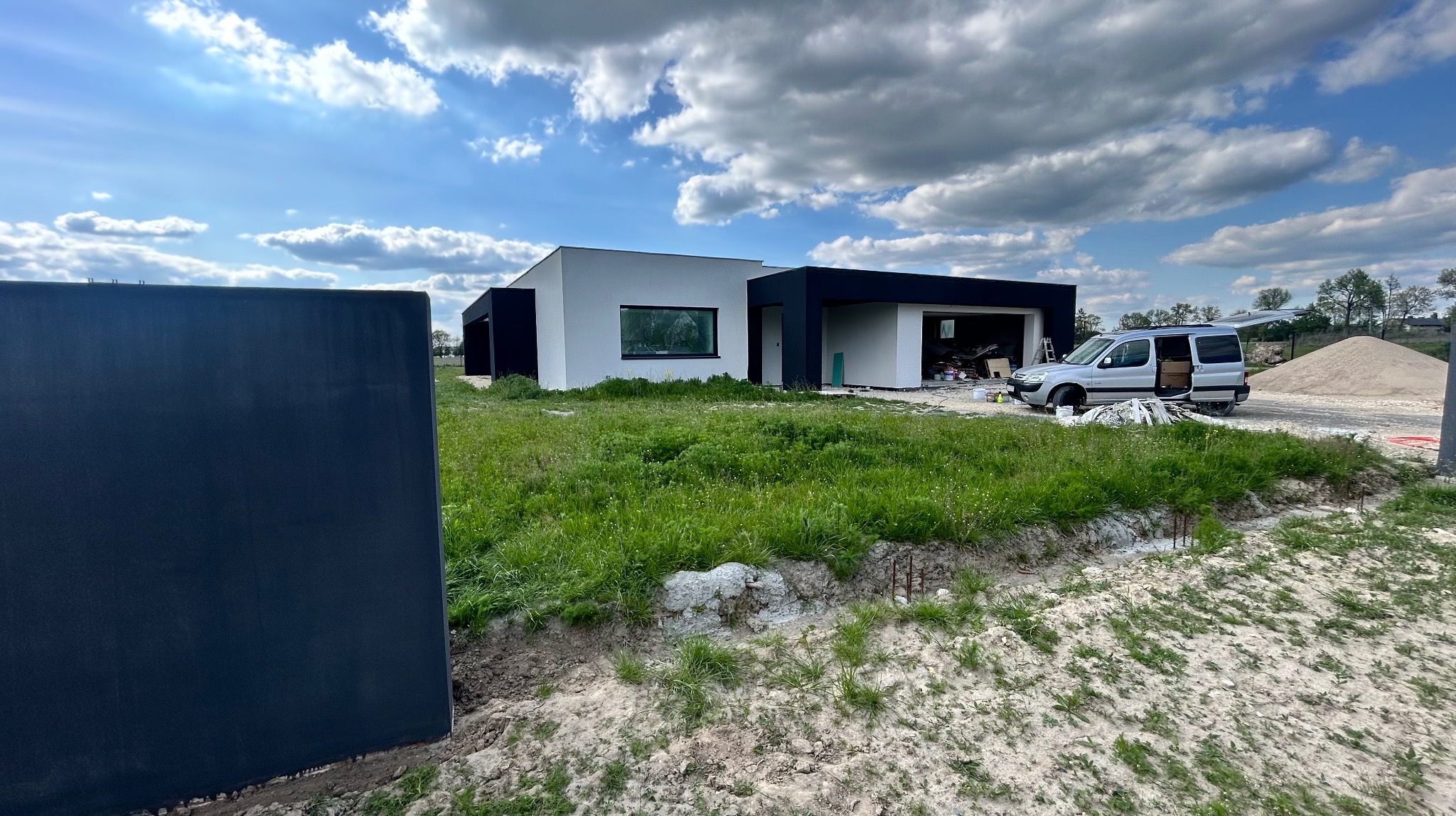 Nowoczesny dom jednorodzinny w trakcie wykończenia, z widocznym samochodem dostawczym i stertą piasku. Częściowo wykończona elewacja i otoczenie domu.