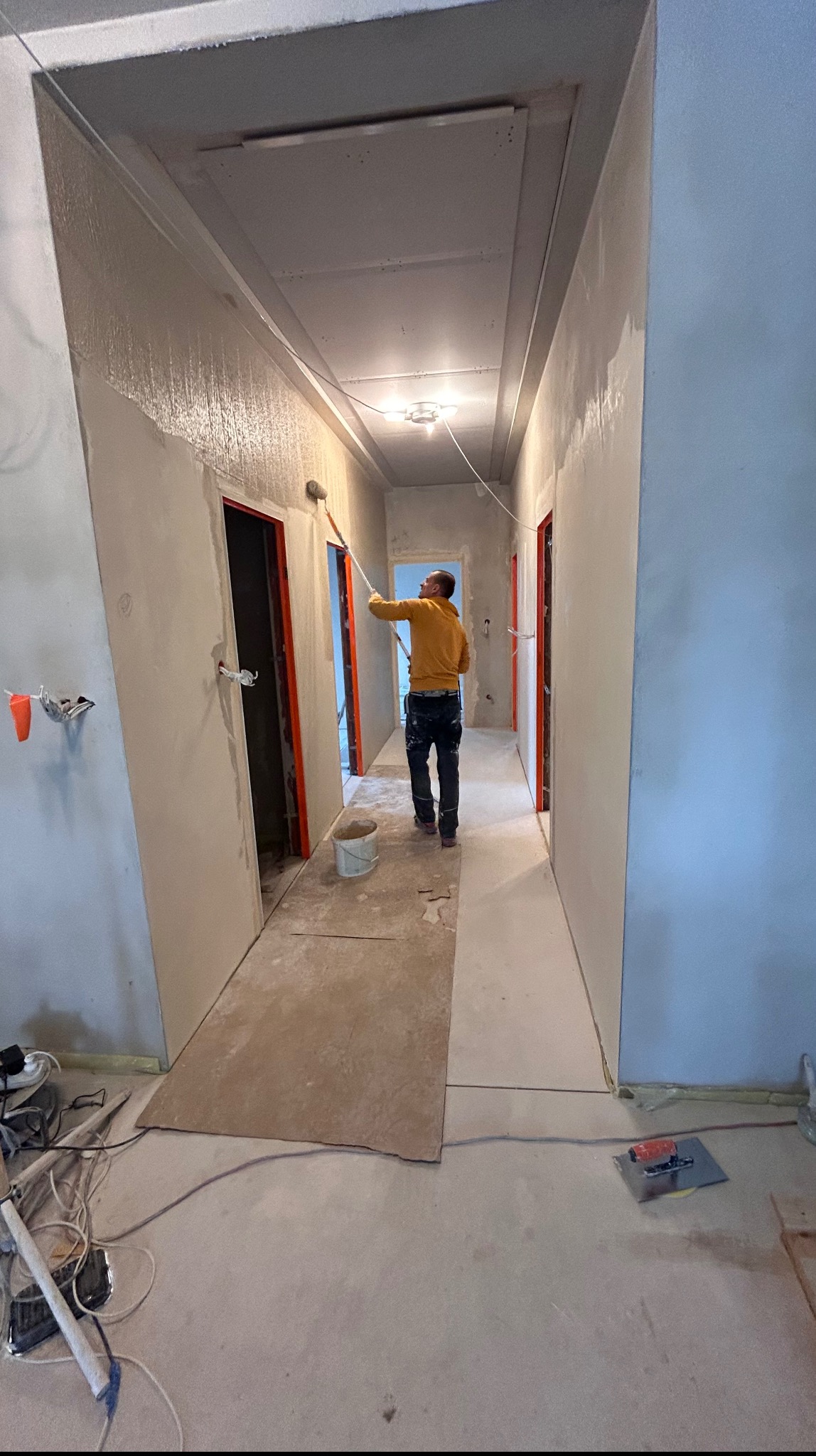 Malowanie wałkiem ścian w korytarzu podczas remontu. Widoczne narzędzia, fragmenty niezabezpieczonej podłogi i pomalowane ściany. Drzwi z pomarańczowymi futrynami.