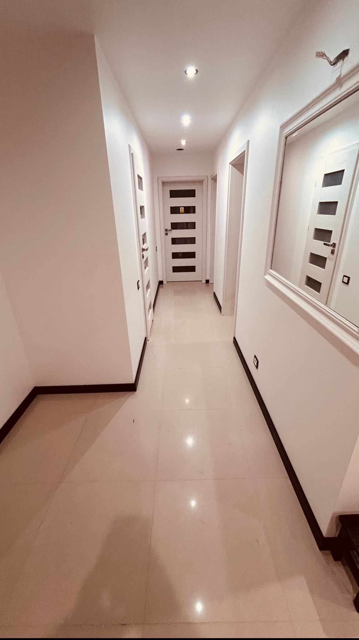 Jasny korytarz po remoncie: białe ściany, kremowa podłoga z płytek, nowe drzwi z przeszkleniami i ozdobne listwy przypodłogowe. Widoczne punkty oświetleniowe w suficie.