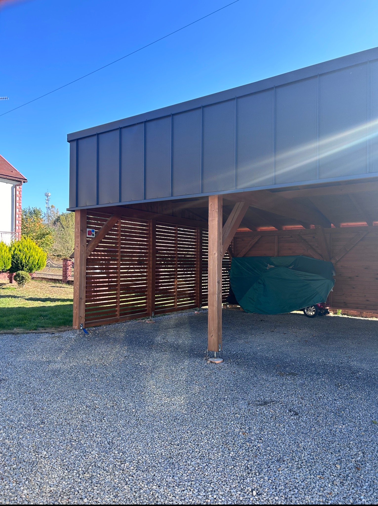 Drewniana wiata garażowa z szarą, metalową konstrukcją dachu, osłonięta ażurowymi panelami, na żwirowym podłożu, w tle dom i zieleń.