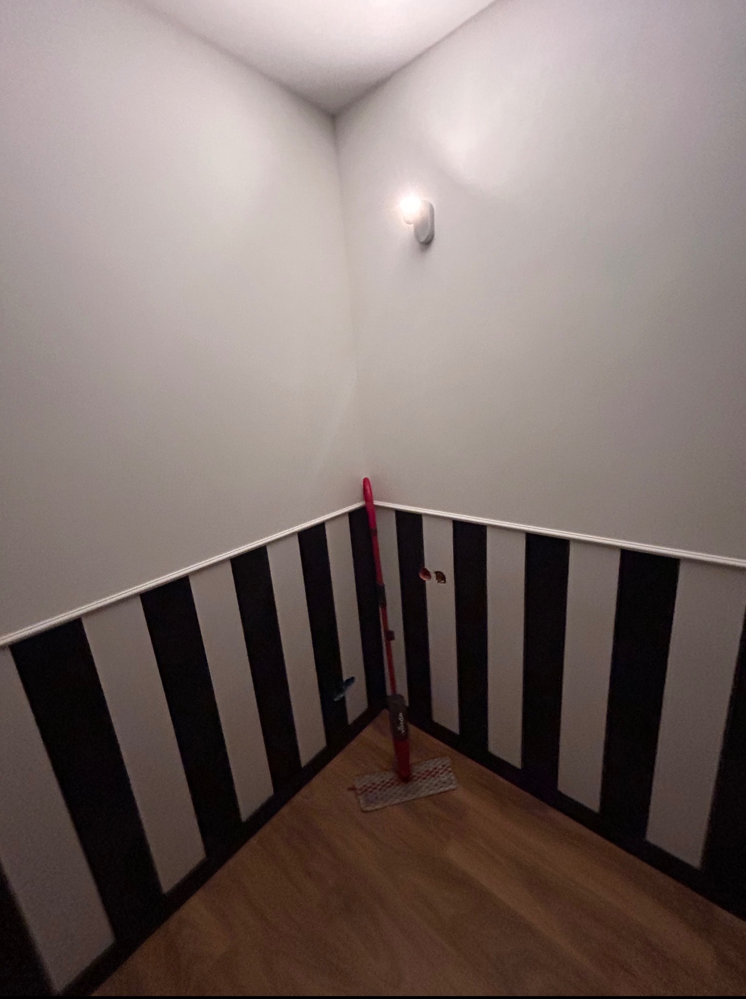 Pokój po remoncie: ściany w czarno-białe pasy, drewniana podłoga. Widoczny mop oparty o ścianę. Oświetlenie punktowe na górze ściany.