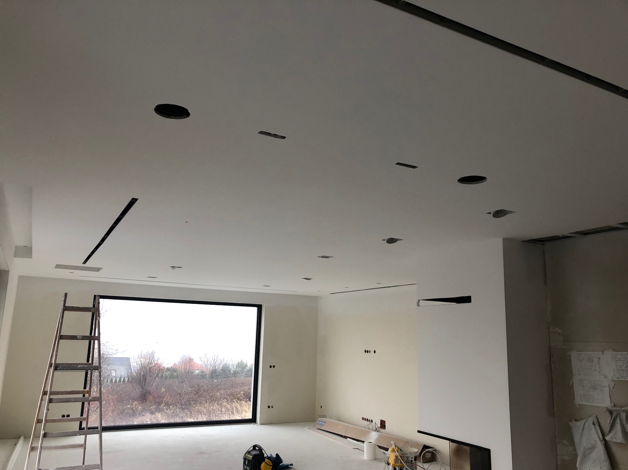 Wnętrze w trakcie wykańczania: białe ściany, sufit z otworami na oświetlenie, drabina przy oknie. Widoczne narzędzia i materiały budowlane.