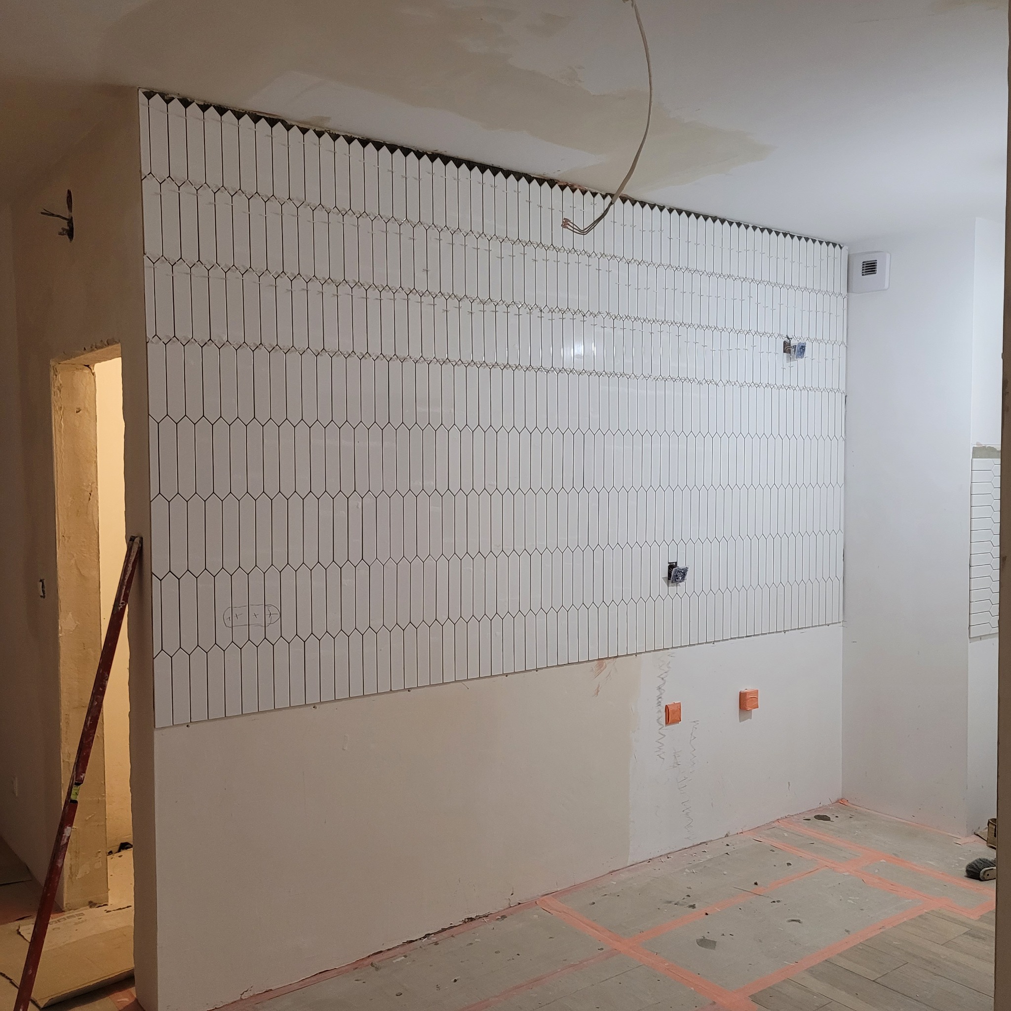 Ściana z białymi, pionowo ułożonymi płytkami w kształcie wydłużonych rombów. Częściowo wykończona ściana z instalacją elektryczną w trakcie prac. Poziomica oparta o ścianę.