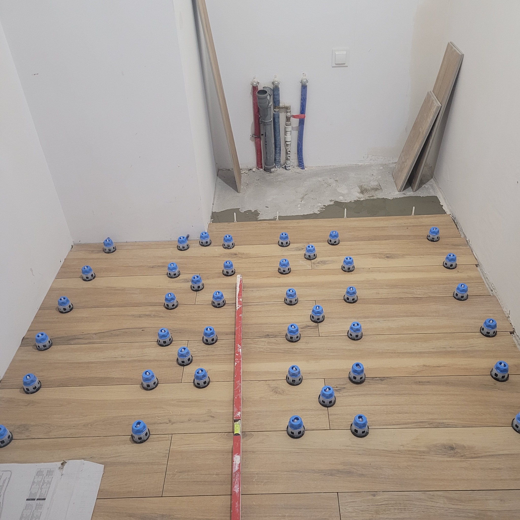 Układanie drewnopodobnych płytek na podłodze łazienki z użyciem systemu poziomowania. Widoczne pionowe rury z instalacją wodną i poziomica.