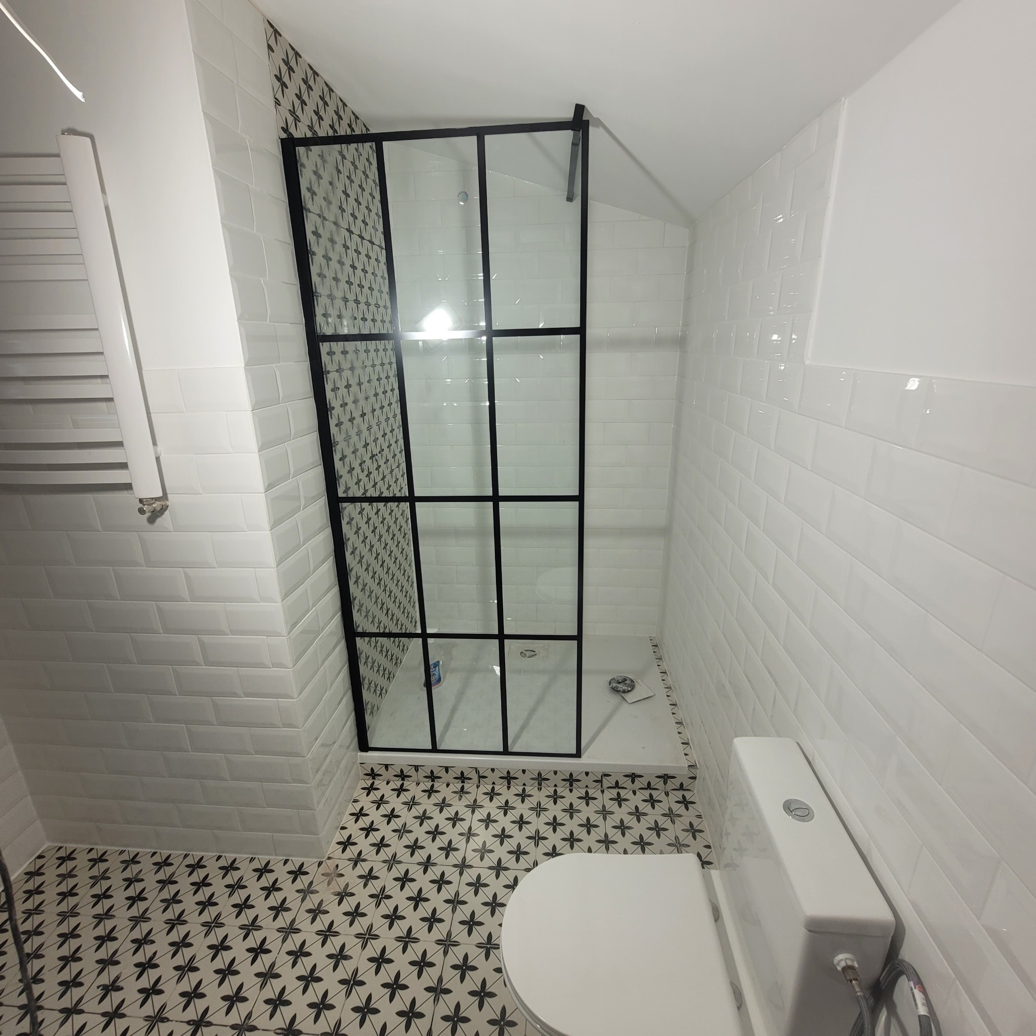 Nowoczesna łazienka z kabiną prysznicową typu walk-in, białe płytki imitujące cegłę na ścianach i wzorzysta podłoga w czarno-białe kwiaty.