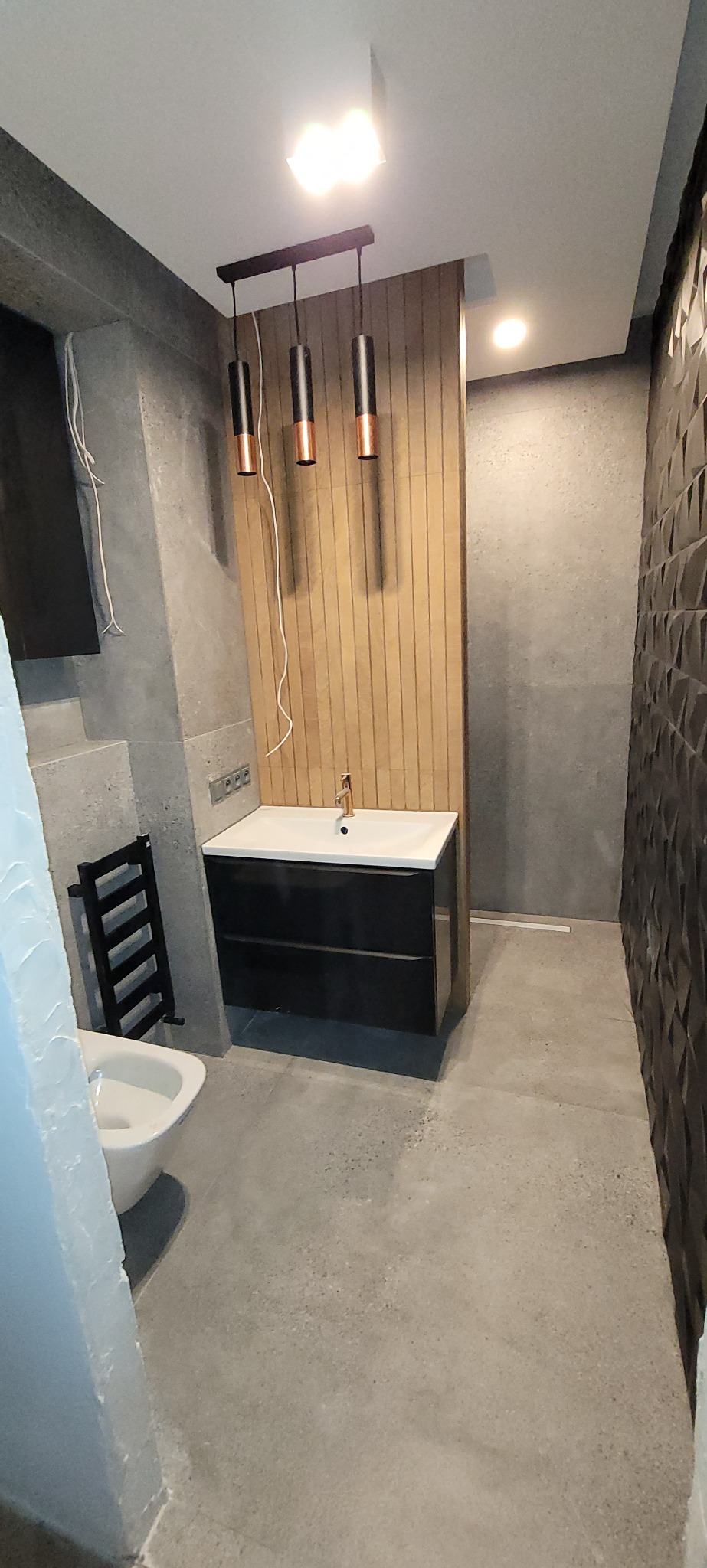 Nowoczesna łazienka w szarościach z akcentami drewna: umywalka z szafką, grzejnik drabinkowy, dekoracyjna ściana z trójwymiarową fakturą i wiszące lampy.