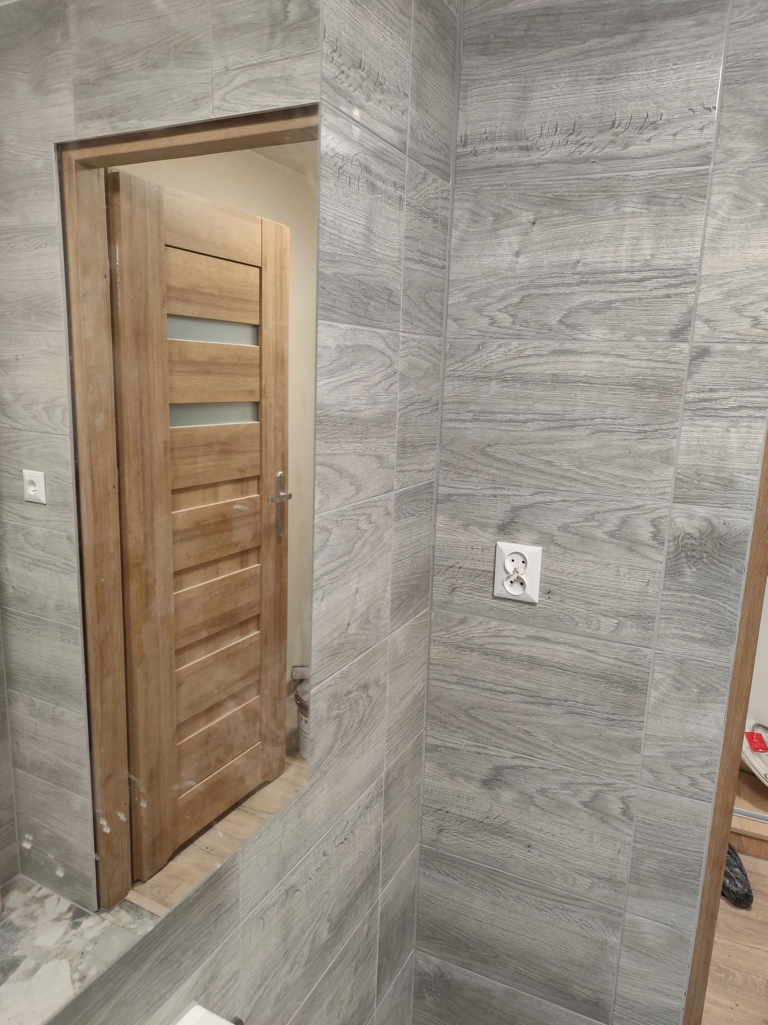 Wykończona łazienka z drewnopodobnymi płytkami i zamontowanymi drewnianymi drzwiami z przeszkleniami. Widoczne gniazdko elektryczne na ścianie.