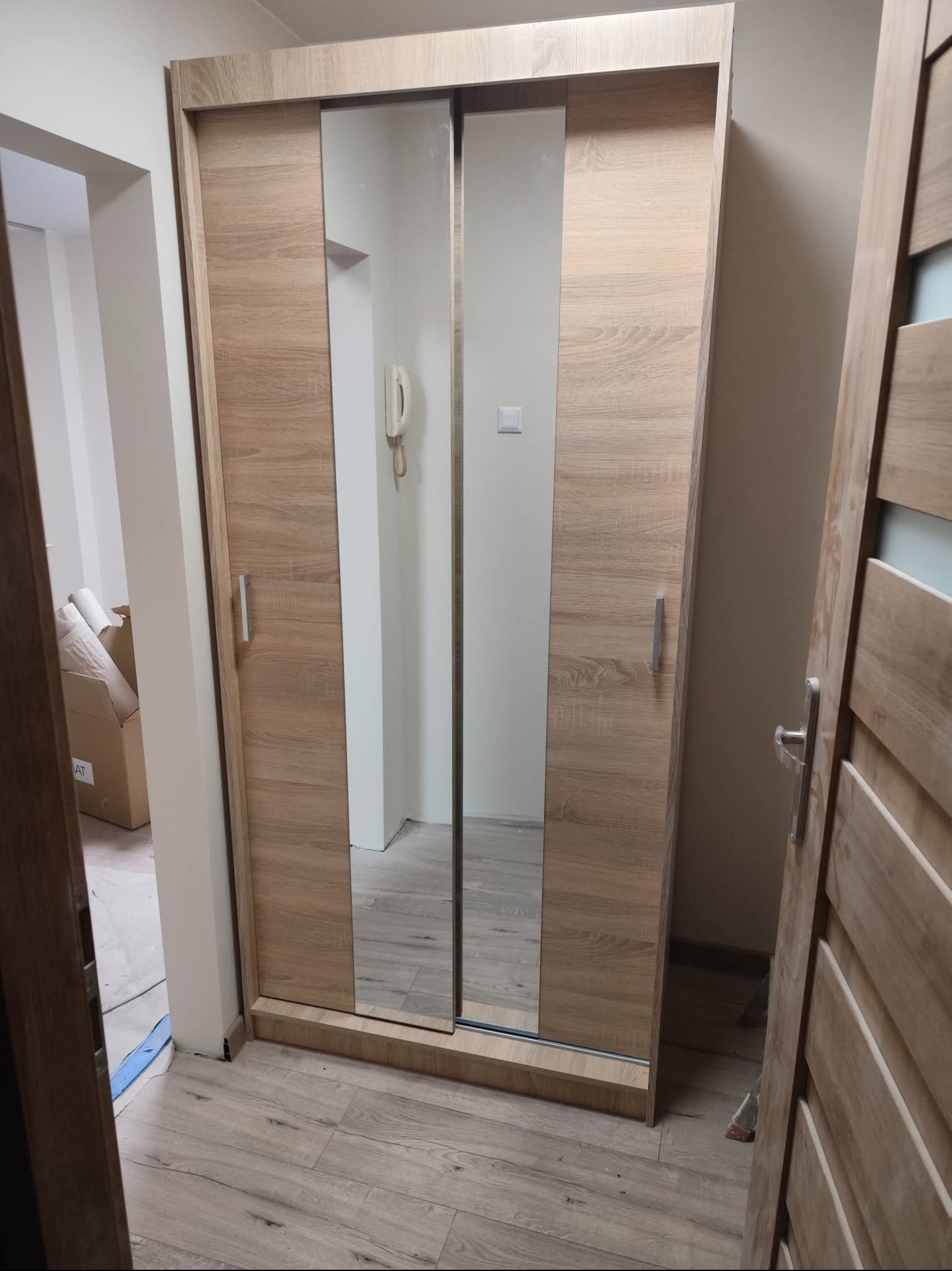 Zamontowana szafa przesuwna z lustrami w przedpokoju, widok z boku. Jasne drewno, minimalistyczne uchwyty, podłoga z paneli. W tle widoczny fragment korytarza.