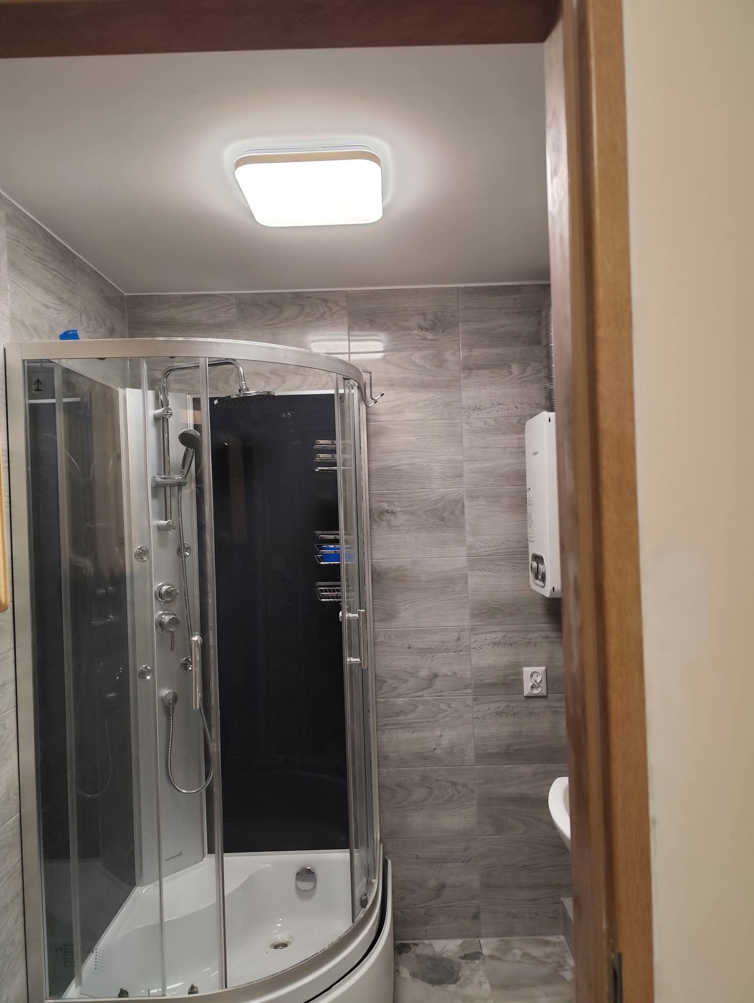 Nowoczesna kabina prysznicowa z hydromasażem, wykończona szarą płytką imitującą drewno. Widoczny bojler i fragment umywalki. Oświetlenie sufitowe.
