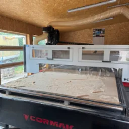 Duża frezarka CNC Cormak w warsztacie, z widocznymi śladami obróbki drewna na stole roboczym. W tle okno i ściany obite płytą OSB.