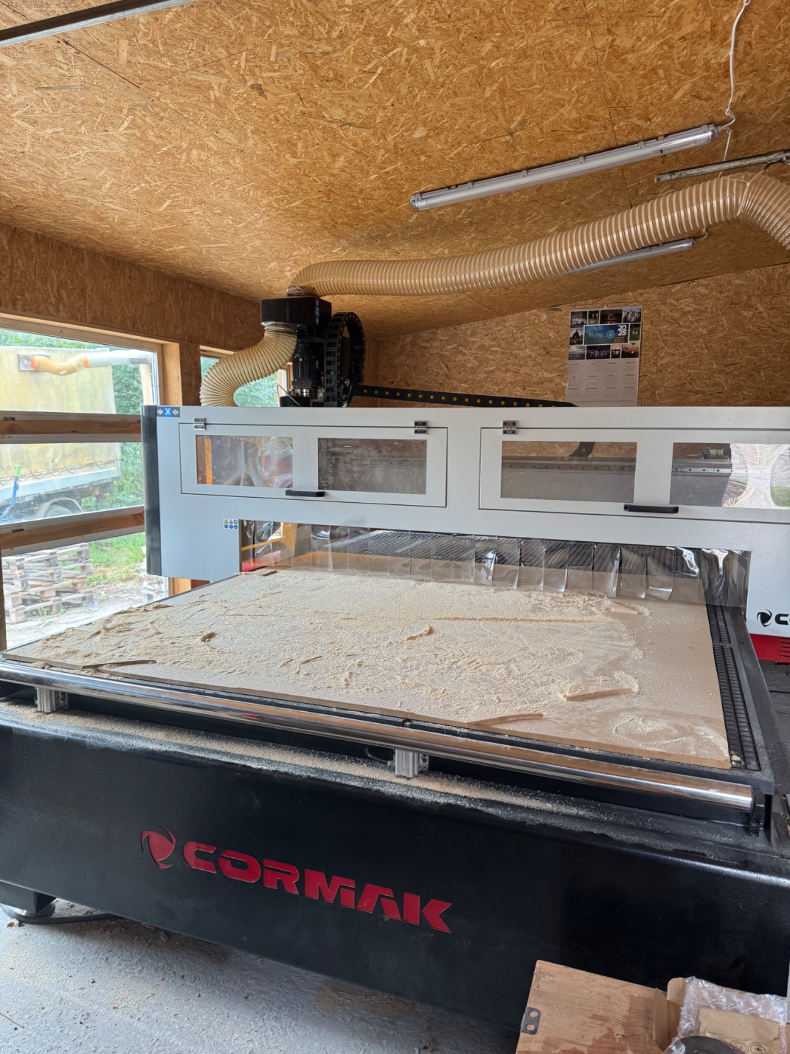 Duża frezarka CNC Cormak w warsztacie, z widocznymi śladami obróbki drewna na stole roboczym. W tle okno i ściany obite płytą OSB.