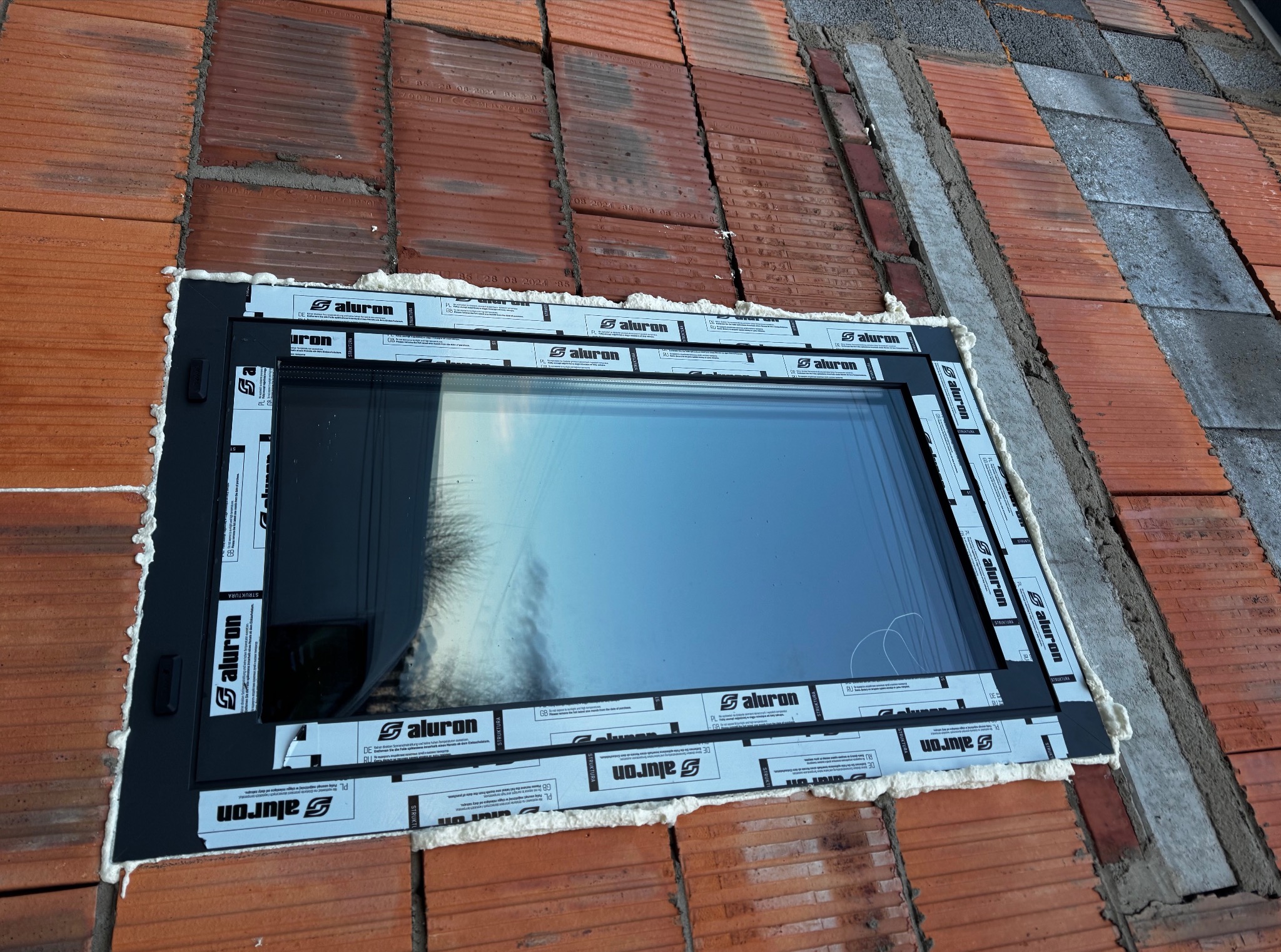 Osadzone okno dachowe w ceglanej konstrukcji dachu, uszczelnione pianą montażową, z widocznymi etykietami producenta na ramie okna.