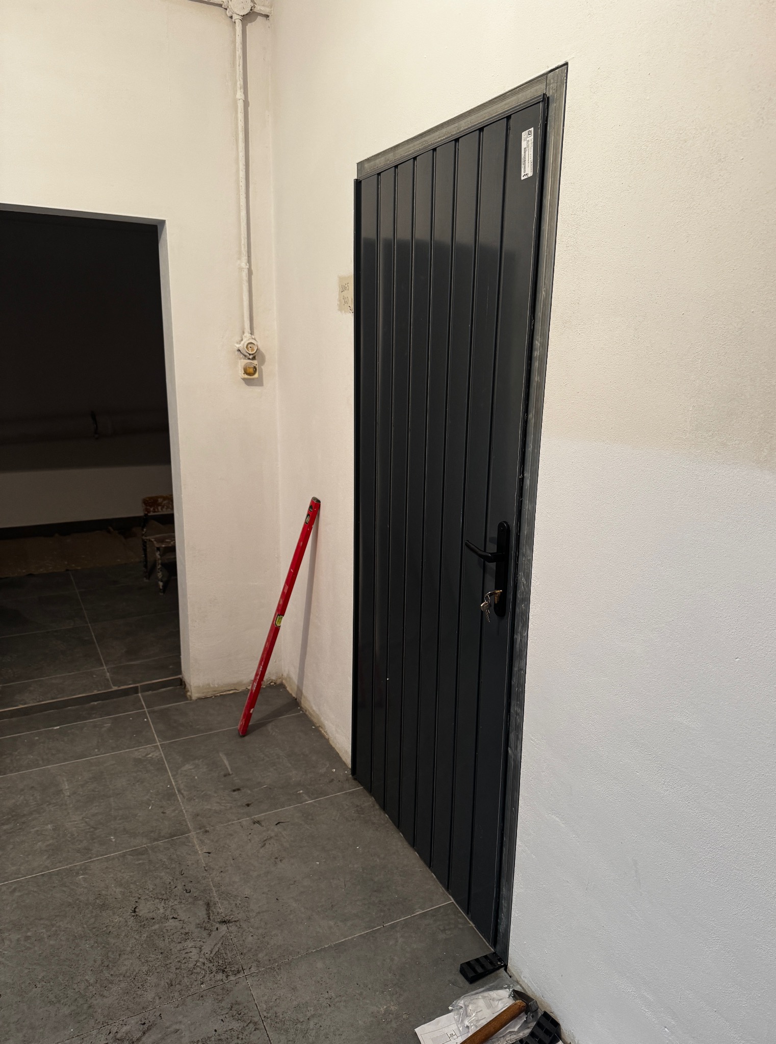 Zamknięte, grafitowe drzwi z pionowymi panelami, zamontowane w białej ścianie. Obok poziomica i narzędzia. Widoczne płytki podłogowe i fragment drugiego pomieszczenia.