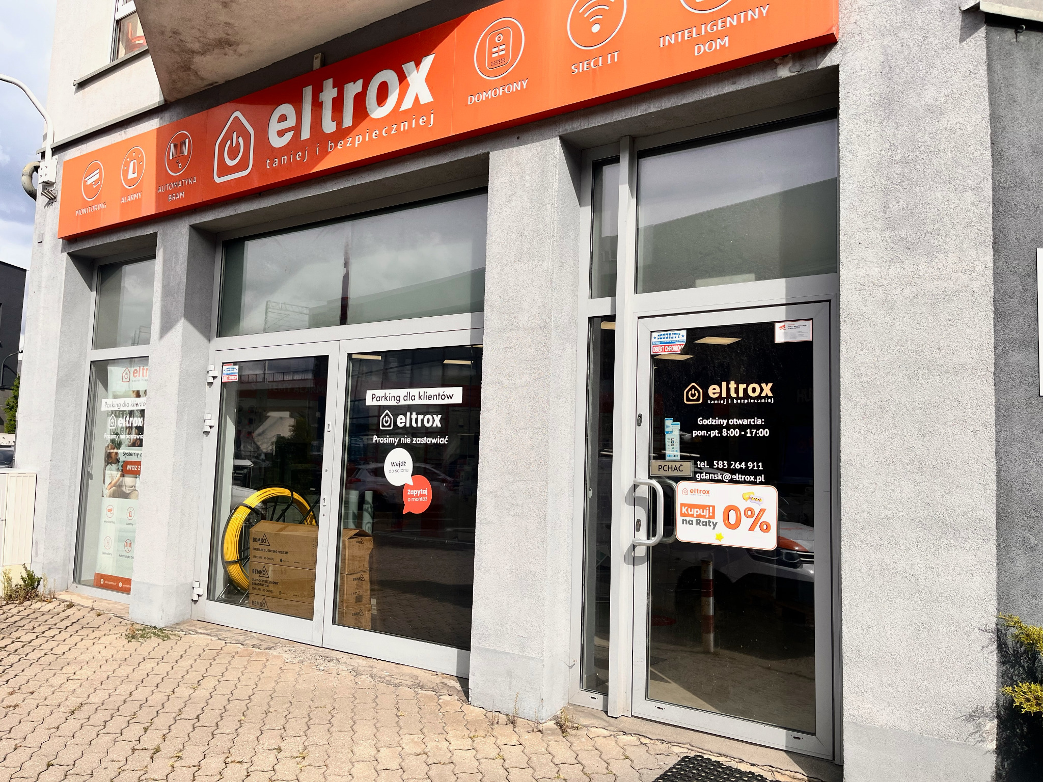 Fasada sklepu Eltrox z ofertą: monitoring, alarmy, automatyka bram, domofony i inteligentny dom. Pomarańczowe logo na szarym budynku. Widoczne reklamy i produkty w oknach.