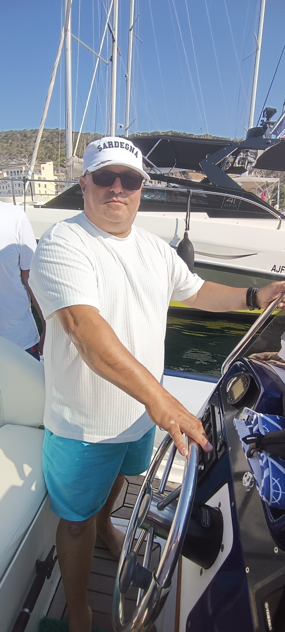 Mężczyzna w czapce 'Sardegna' steruje jachtem motorowym w słoneczny dzień. Widoczny fragment kokpitu z chromowaną kierownicą i panelem sterowania.