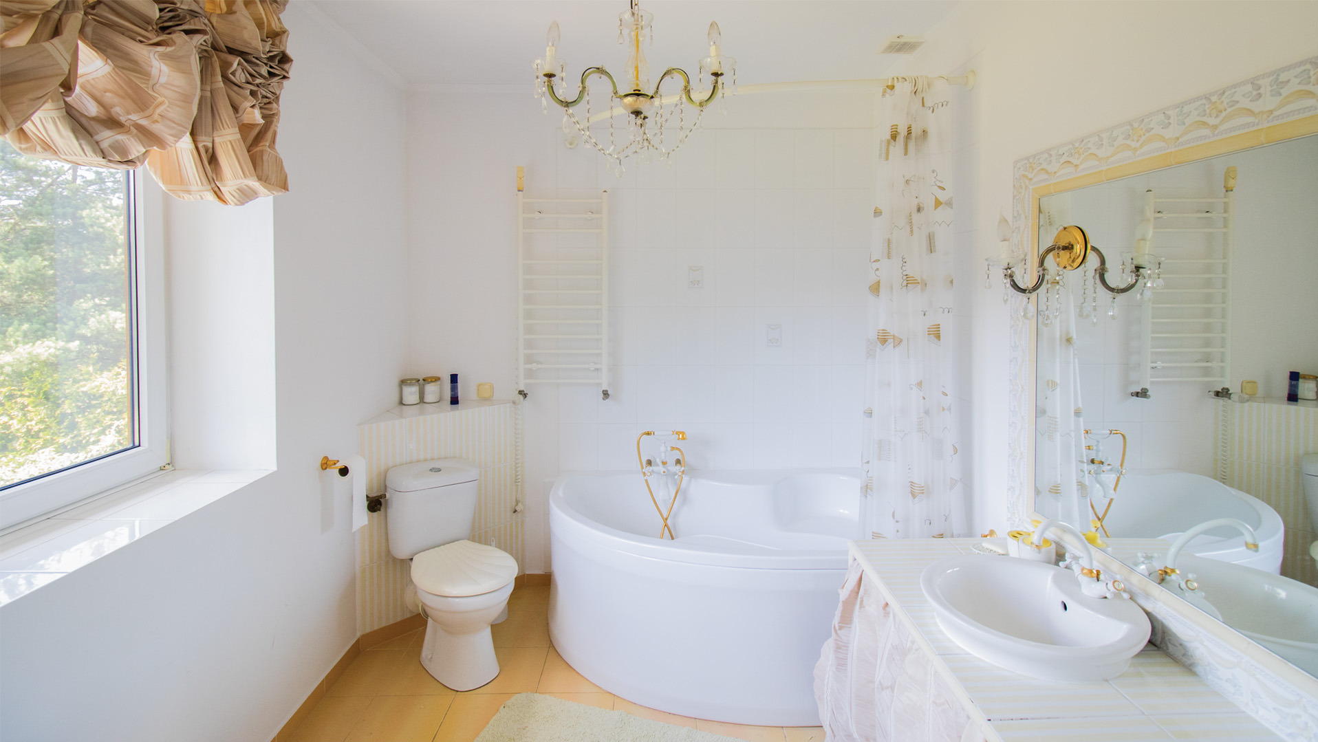 Jasna łazienka z wanną narożną, toaletą i oknem. Kryształowy żyrandol dodaje elegancji. Uwagę zwraca złota armatura i dekoracyjne zasłony przy oknie.