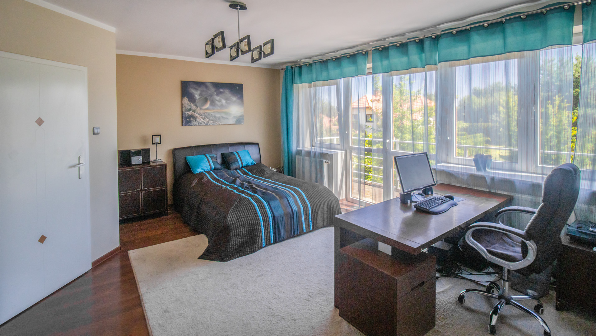 Jasna sypialnia z biurkiem przy oknie, łóżkiem z turkusowymi akcentami i ciemnymi meblami. Widok na zieleń za oknem. Nowoczesne oświetlenie sufitowe.