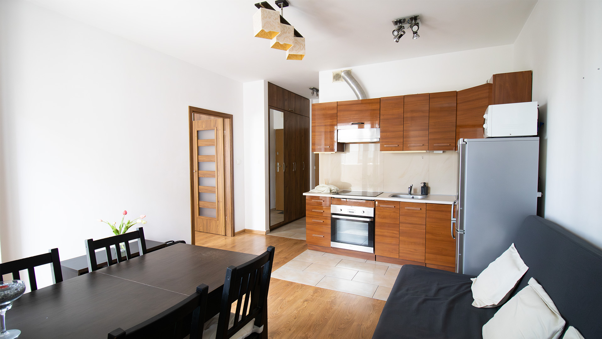 Jasne wnętrze mieszkania w Grodzisku Mazowieckim: stół z krzesłami, aneks kuchenny z brązowymi szafkami, szara lodówka i sofa. Widok na drzwi i szafę.