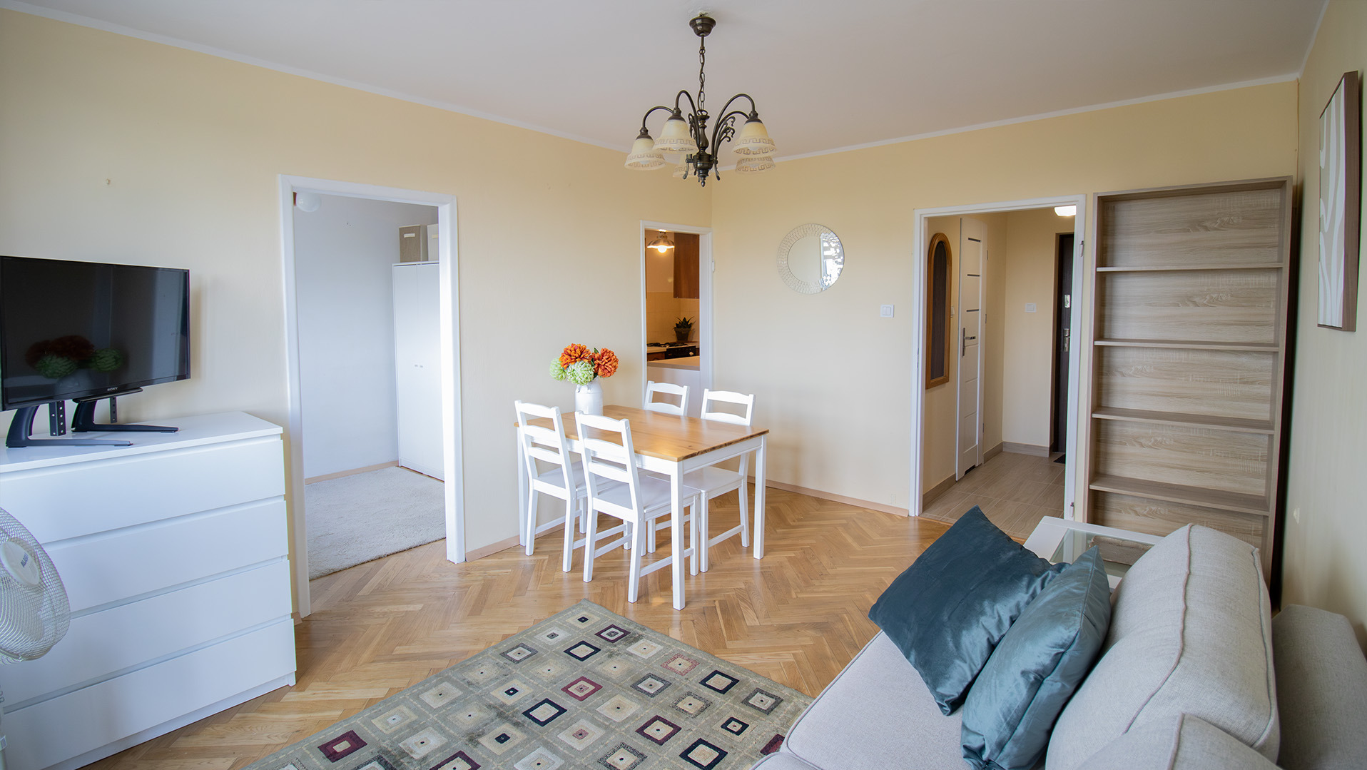 Jasne wnętrze salonu z parkietem w jodełkę. Widoczny stół z krzesłami, kanapa z poduszkami, regał i telewizor. W tle otwarte drzwi do innych pomieszczeń.