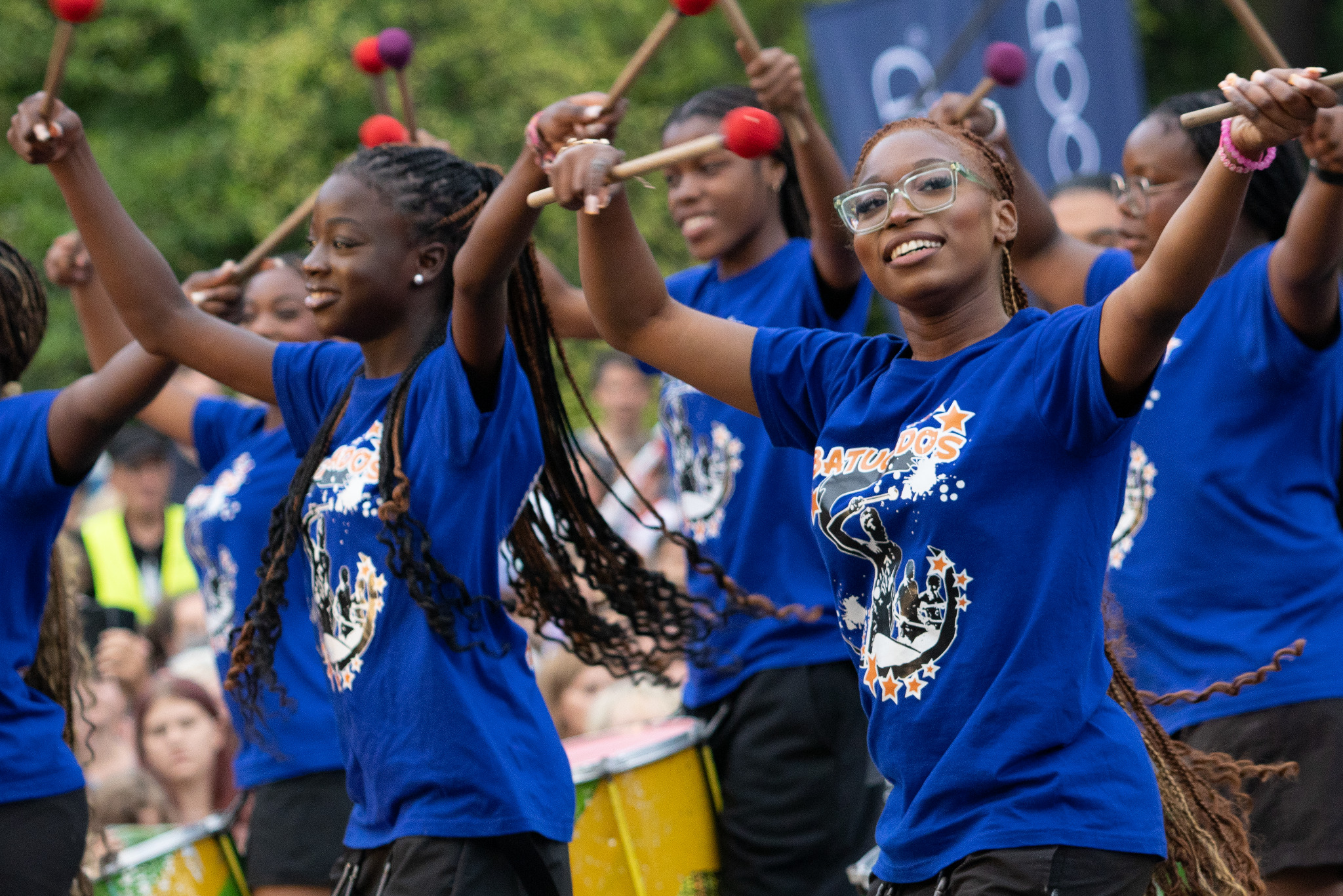 Zespół perkusyjny w niebieskich koszulkach gra na bębnach podczas występu. Uśmiechnięta kobieta w okularach na pierwszym planie, w tle publiczność.