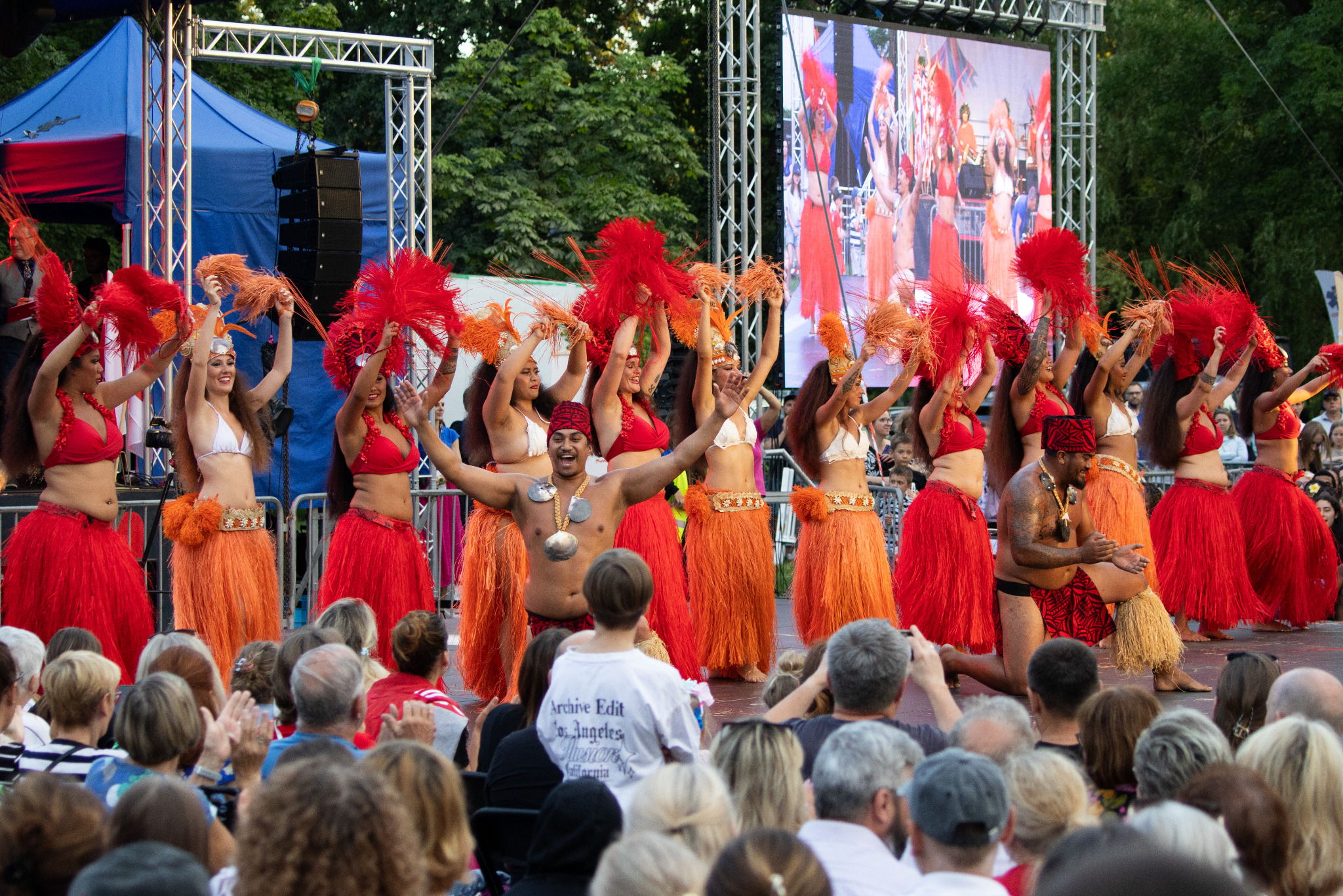 Barwny występ taneczny na scenie plenerowej, z tancerzami w tradycyjnych strojach i publicznością. Scena z ekranem LED w tle, część tancerzy z rekwizytami.