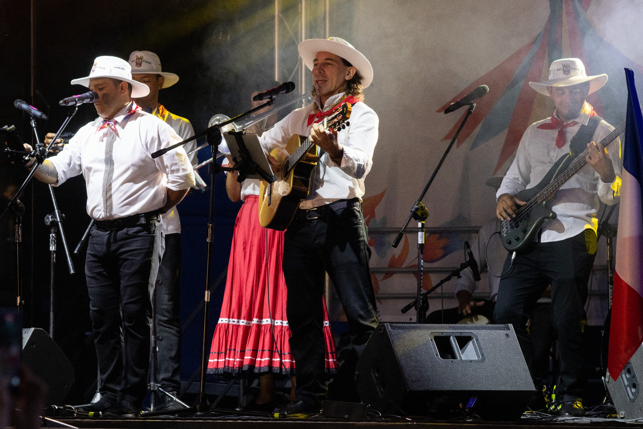 Zespół muzyczny w strojach ludowych na scenie podczas występu. Wokalista gra na gitarze, a pozostali muzycy akompaniują na instrumentach.