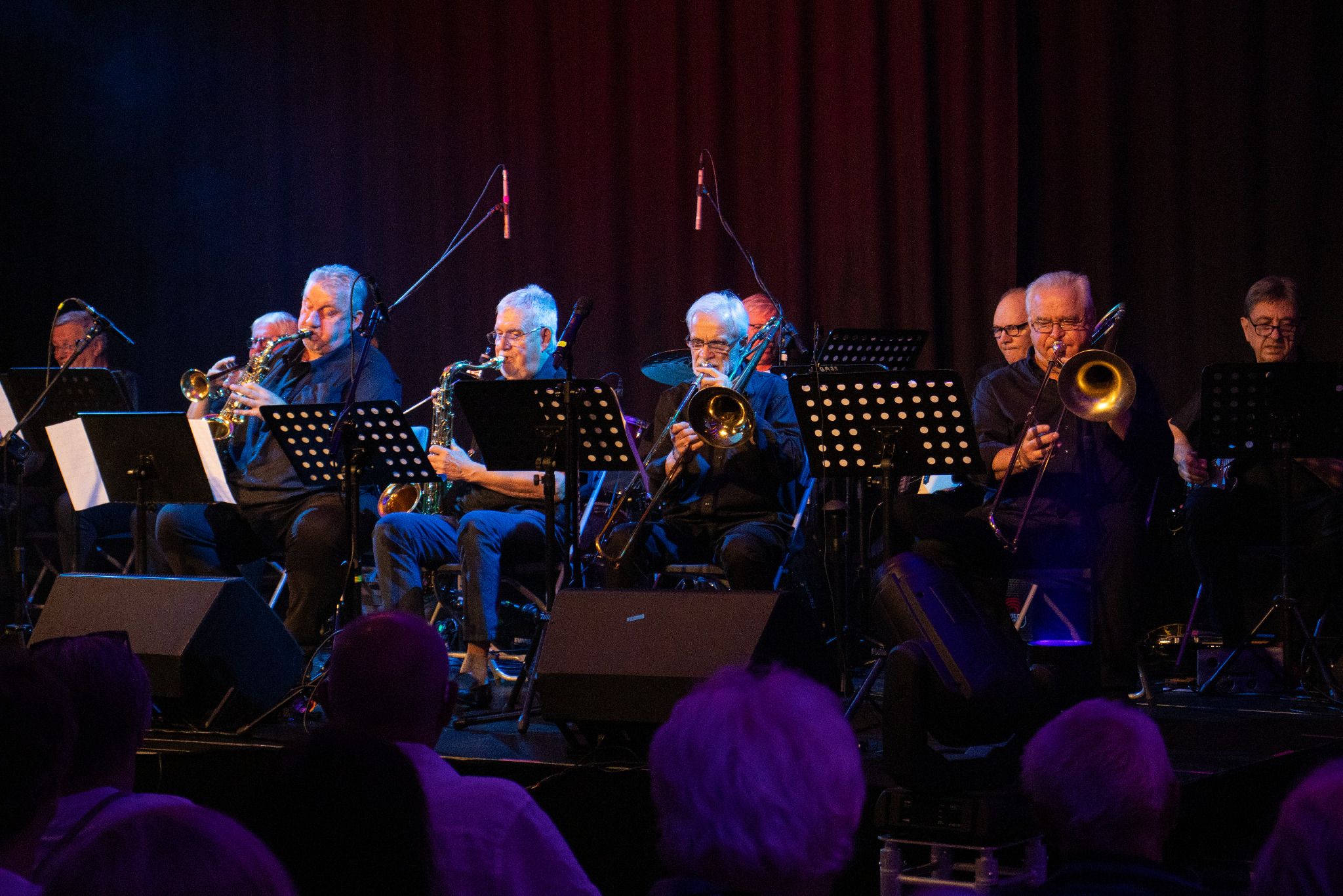 Zespół muzyczny na scenie podczas występu. Widoczni muzycy grający na saksofonach i puzonach, oświetleni kolorowym światłem. Widownia w tle.