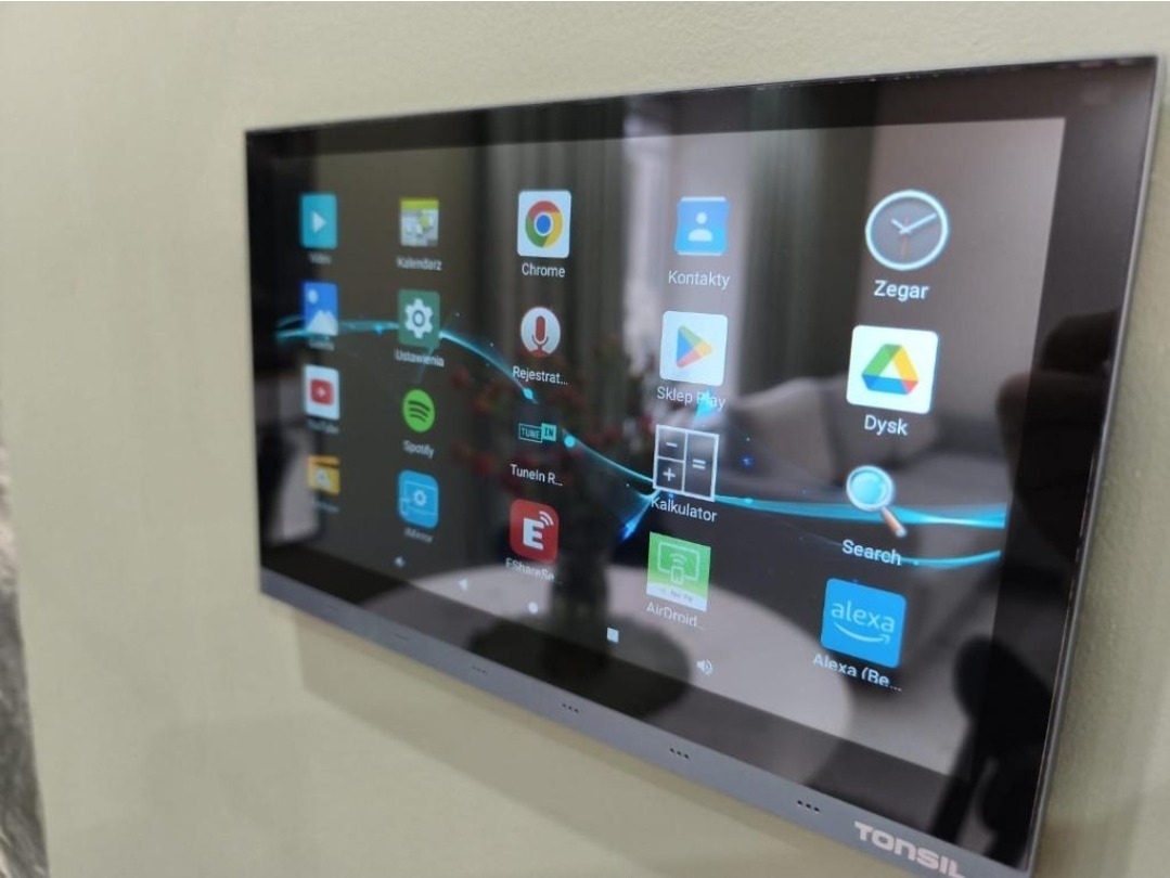 Panel dotykowy Tonsil zamontowany na ścianie, wyświetlający ekran główny z ikonami aplikacji. Widok z boku, ukazujący detale montażu.