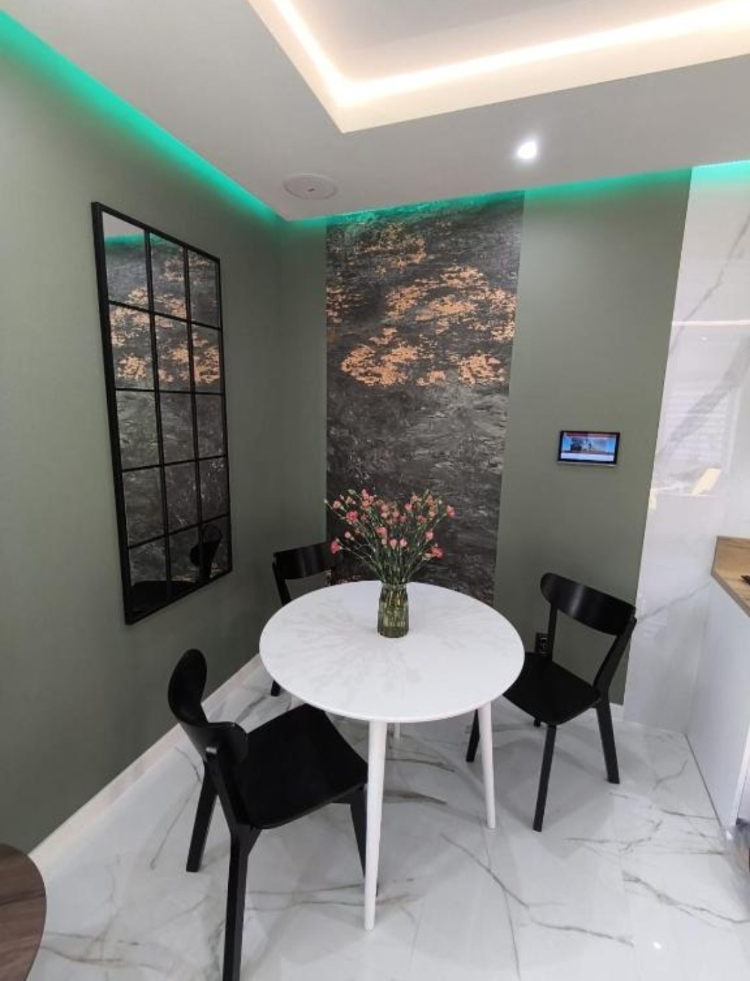 Nowoczesna jadalnia z okrągłym białym stołem i czarnymi krzesłami na marmurowej podłodze. Dekoracyjna ściana, lustro z czarną ramą i zielone oświetlenie LED.