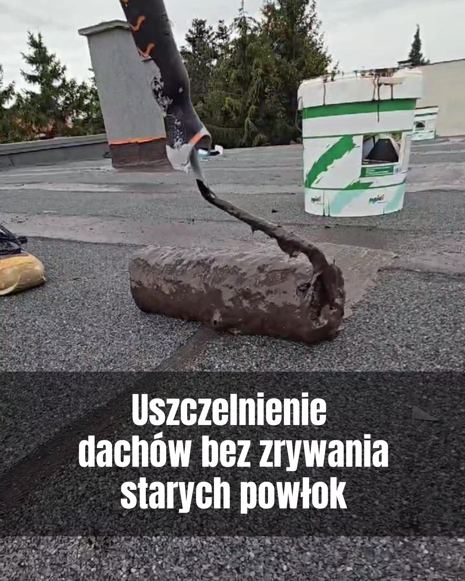 Wałek malarski aplikuje hydroizolację na dach pokryty papą. W tle widoczne wiadro z preparatem i komin wentylacyjny. Praca wykonywana na zewnątrz.