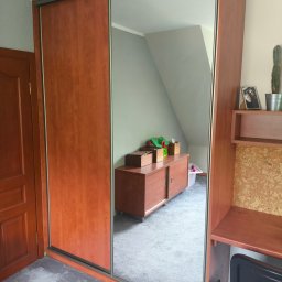 ULMAN - Brązowa szafa przesuwna z lustrem w pokoju na poddaszu. W lustrze odbija się komoda i biurko. Widoczny fragment drzwi i dywanu. Całość w ciepłej tonacji.
