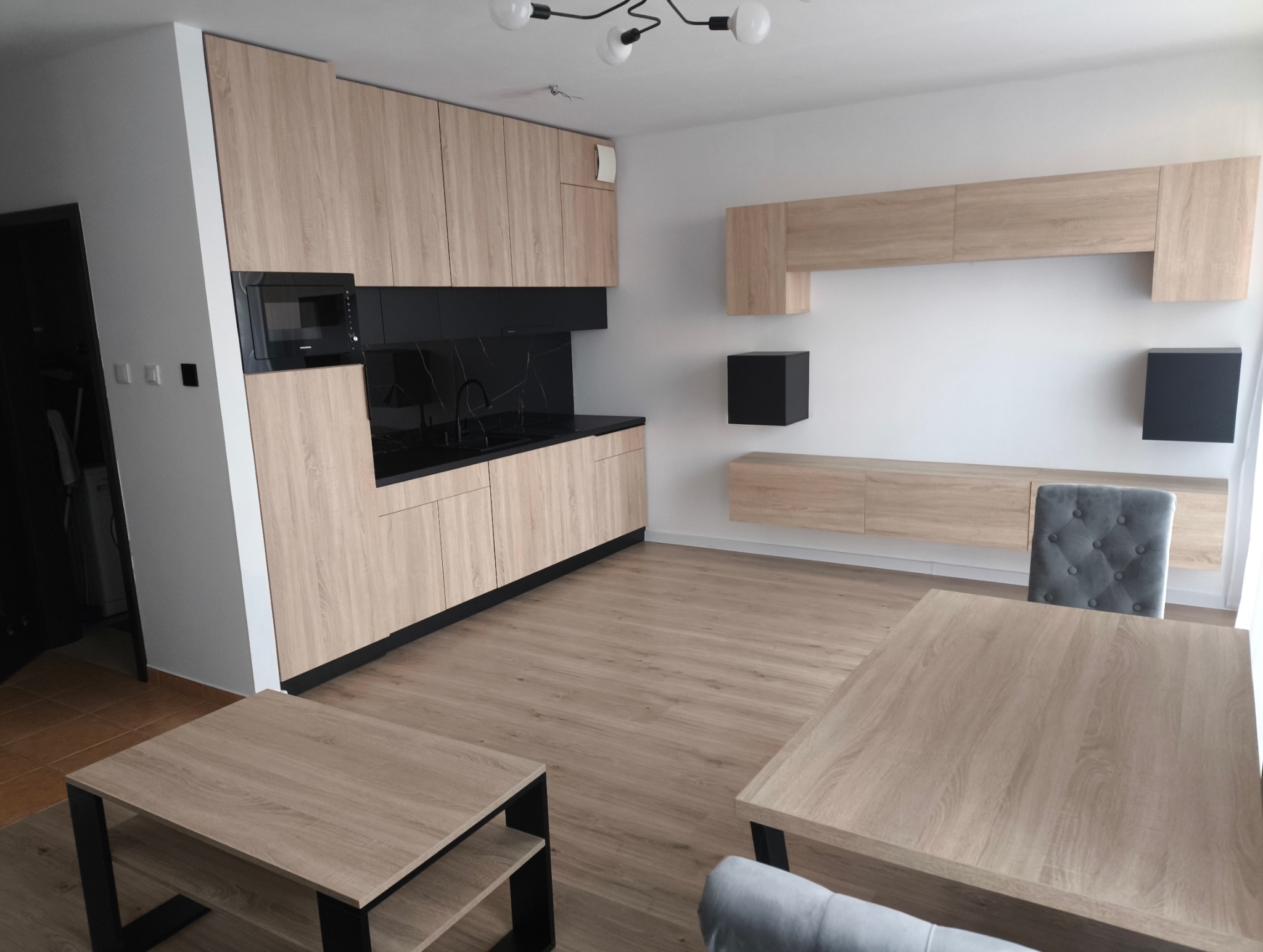 Nowoczesna aranżacja wnętrza: kuchnia z zabudową w odcieniach drewna i czarnym blatem, panele podłogowe w ciepłym odcieniu, stół i krzesła w stonowanej kolorystyce.