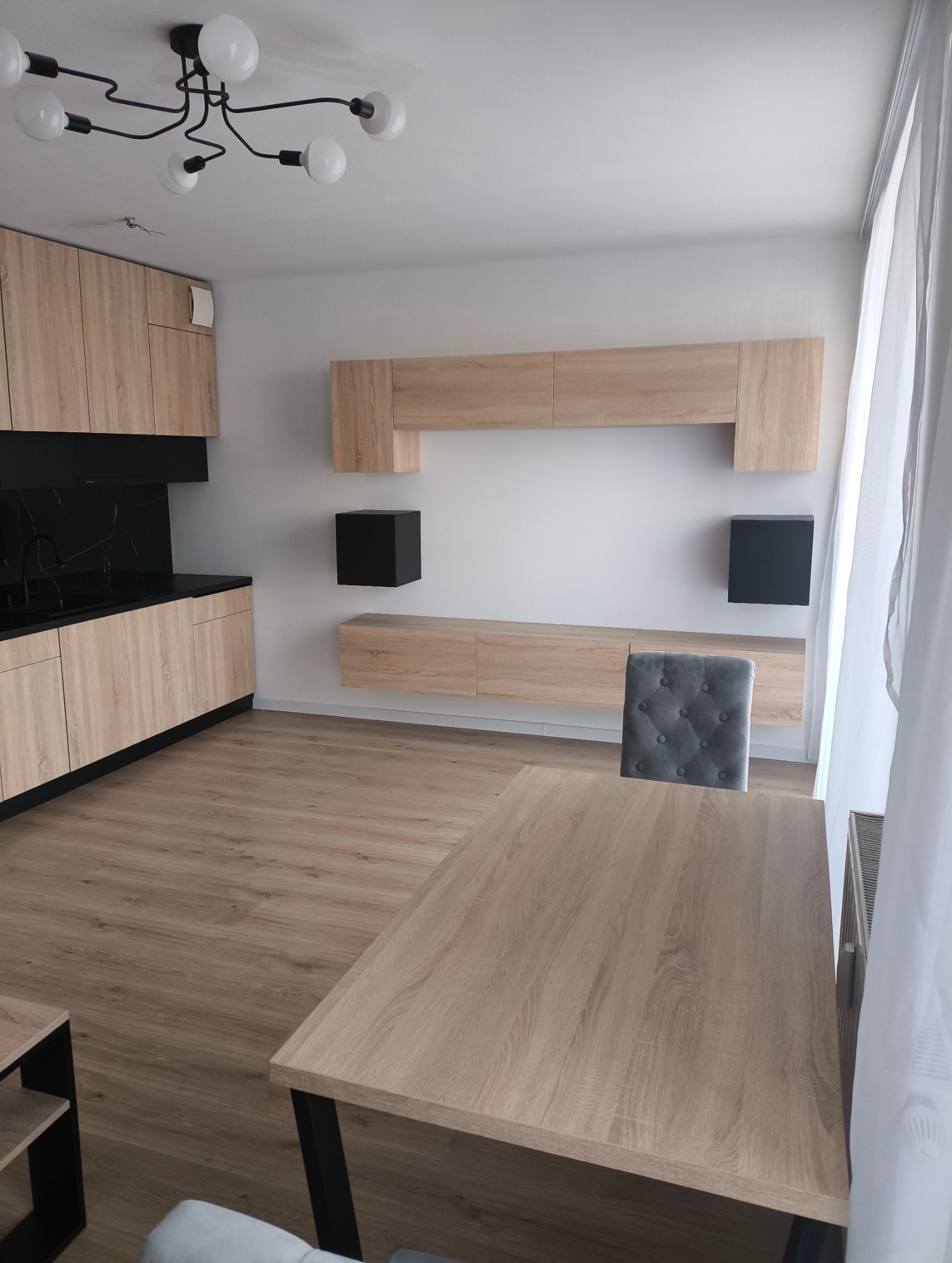 Jasne panele podłogowe, stół z czarnymi nogami i szare krzesło w pokoju z zabudową meblową w odcieniach drewna i czarnymi akcentami, nowoczesne oświetlenie sufitowe.
