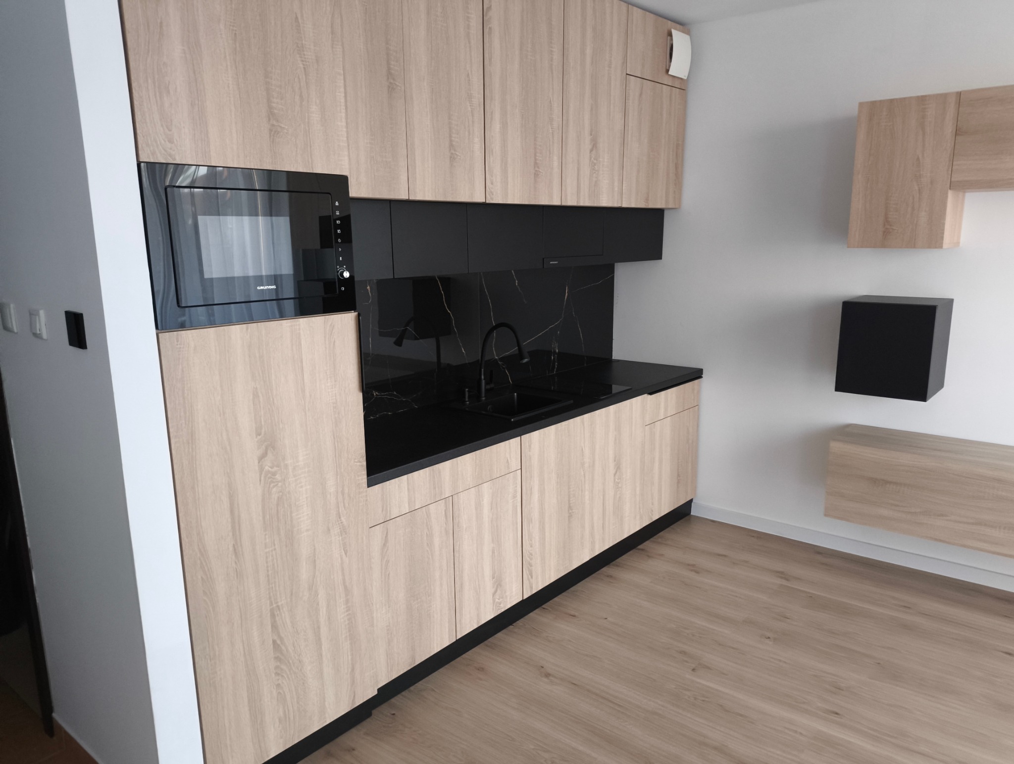 Jasna, nowoczesna kuchnia z drewnianymi frontami i czarnym blatem. Widoczna zabudowa z mikrofalówką i szafkami wiszącymi, minimalistyczne dodatki na ścianie.