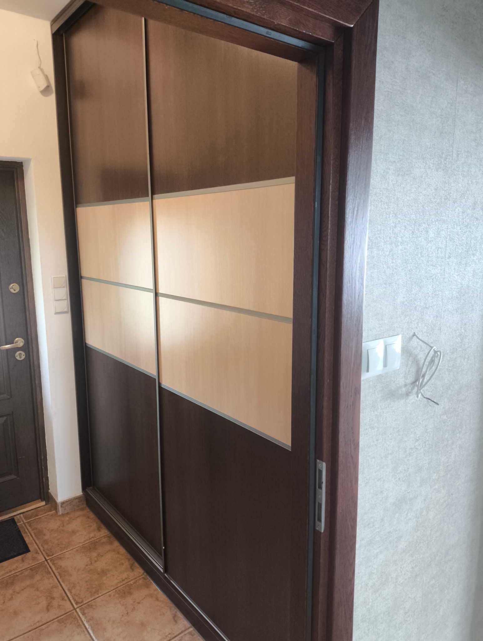 Dwukolorowa szafa wnękowa z przesuwnymi drzwiami, brązowo-beżowa, z widocznym fragmentem korytarza i brązowymi płytkami podłogowymi.