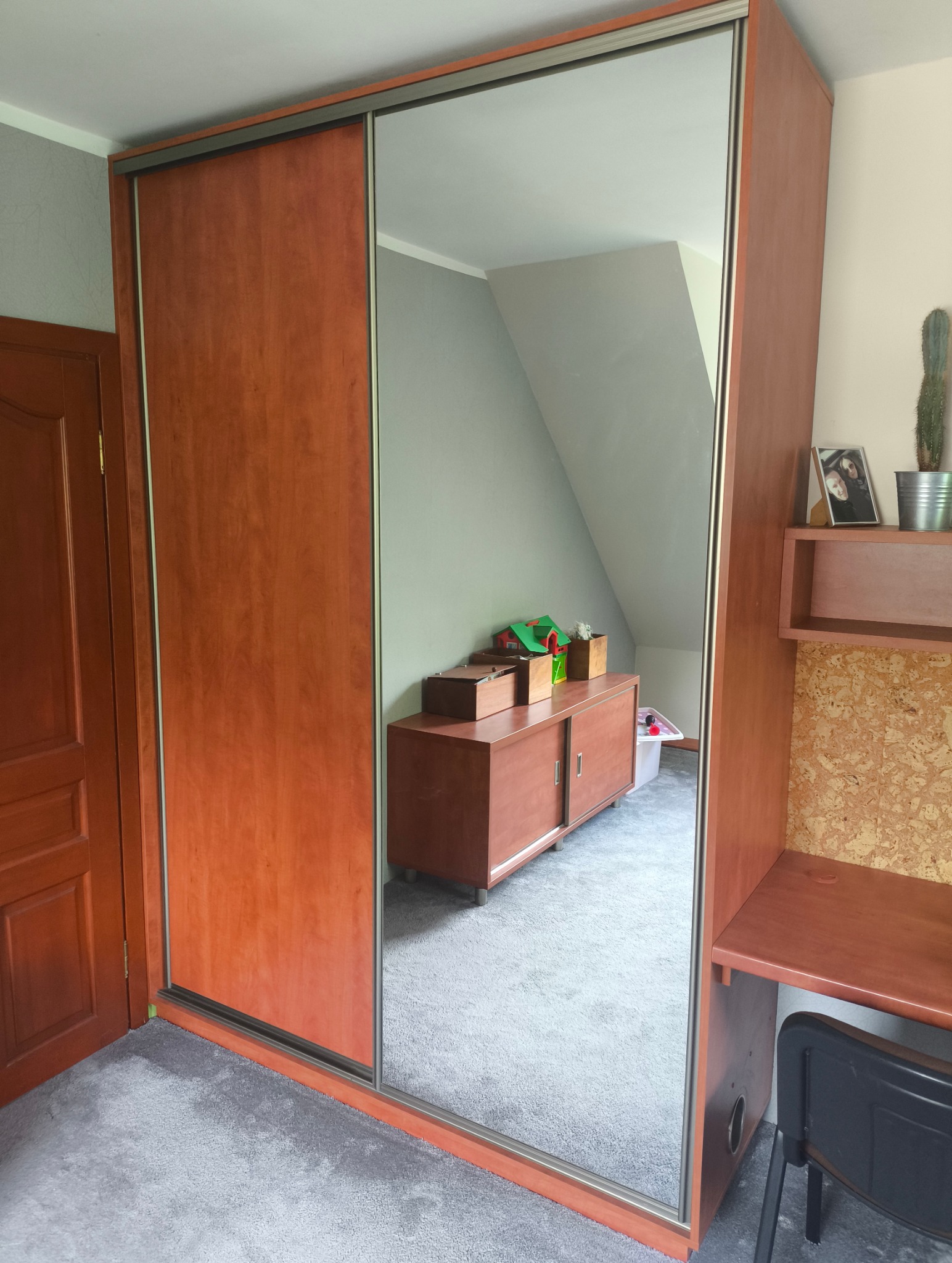 Brązowa szafa przesuwna z lustrem w pokoju na poddaszu. W lustrze odbija się komoda i biurko. Widoczny fragment drzwi i dywanu. Całość w ciepłej tonacji.