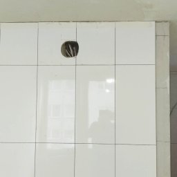 Jacek Woźny - Ściana wyłożona białymi, błyszczącymi płytkami. Widoczny otwór na gniazdko elektryczne z wystającymi kablami. W odbiciu na płytkach widoczna sylwetka osoby.