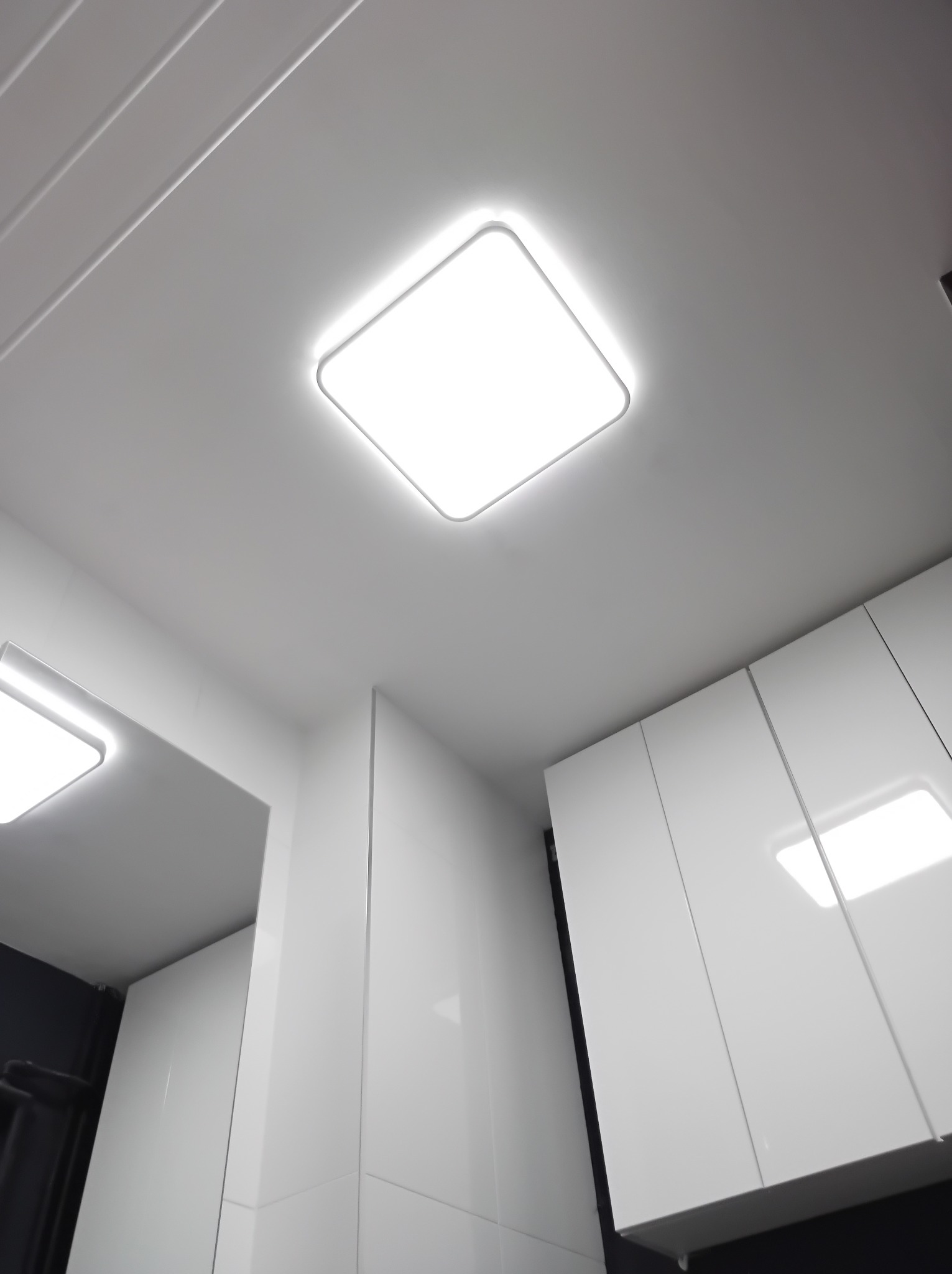 Nowoczesna łazienka z białymi, błyszczącymi szafkami pod sufitem i kwadratową lampą LED w zabudowie. Minimalistyczny design, czyste linie, oświetlenie sufitowe.