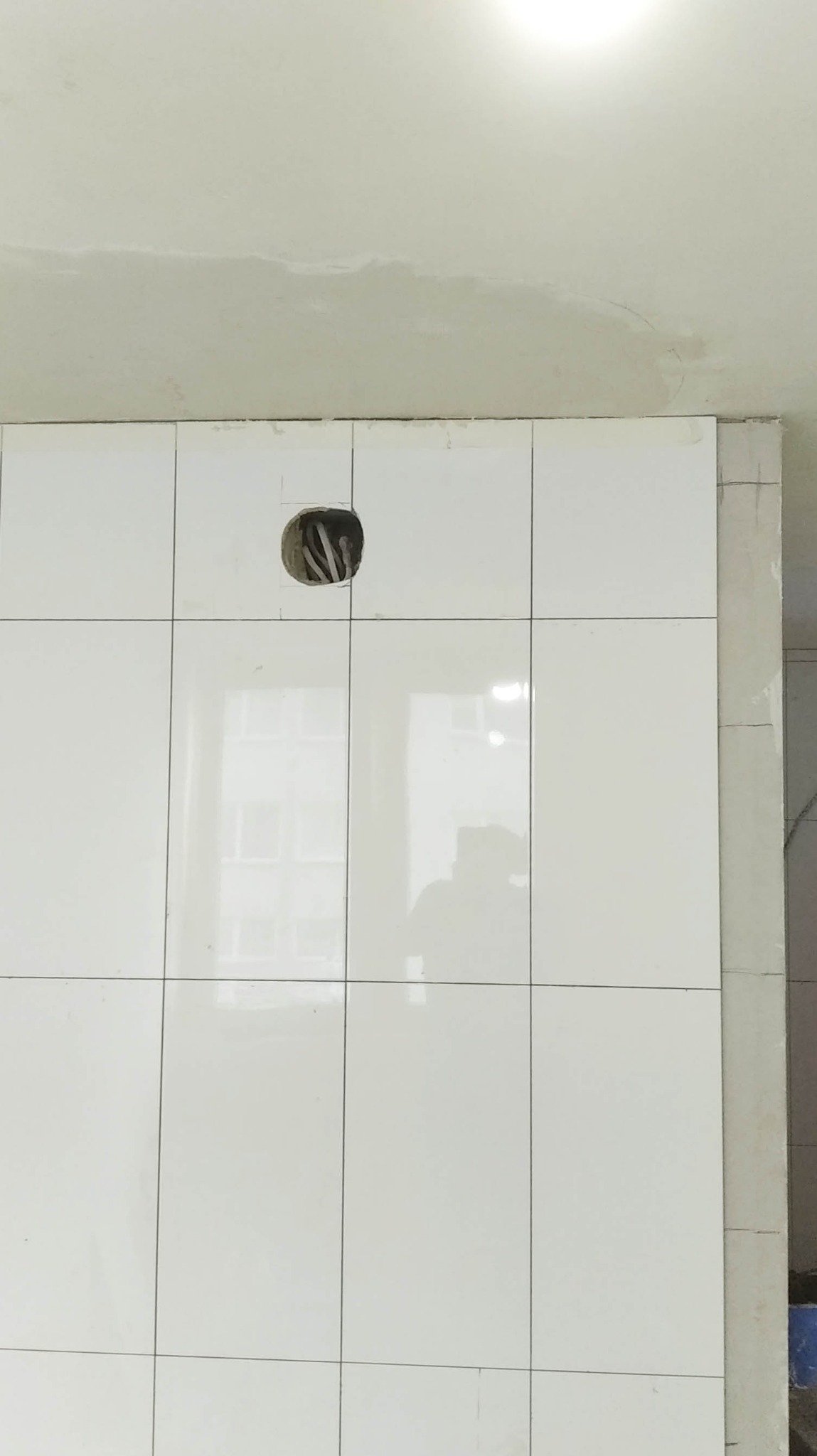 Ściana wyłożona białymi, błyszczącymi płytkami. Widoczny otwór na gniazdko elektryczne z wystającymi kablami. W odbiciu na płytkach widoczna sylwetka osoby.