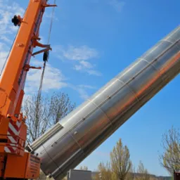 Pomarańczowy dźwig podnosi metalowy, cylindryczny element na tle błękitnego nieba. Pracownik w kamizelce odblaskowej obserwuje operację.