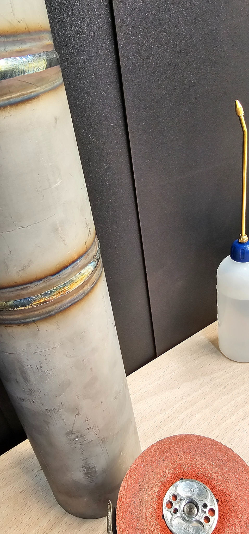 Metalowa rura ze spoiną, obok narzędzia szlifierskie i butelka z płynem, na jasnym blacie i czarnym tle, ukazująca etap obróbki metalu.