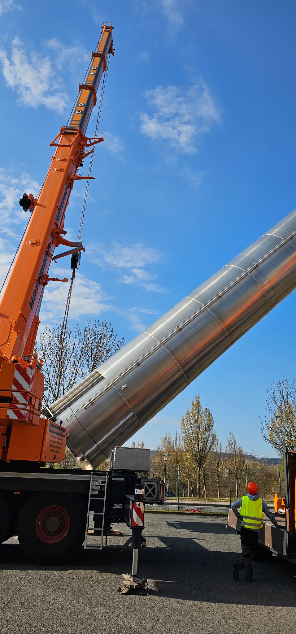 Pomarańczowy dźwig podnosi metalowy, cylindryczny element na tle błękitnego nieba. Pracownik w kamizelce odblaskowej obserwuje operację.