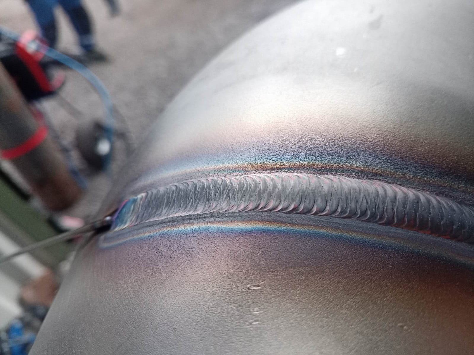 Spoina na metalowej rurze z charakterystycznymi tęczowymi przebarwieniami, widoczne ślady obróbki. Detal spoiny wykonanej metodą TIG.