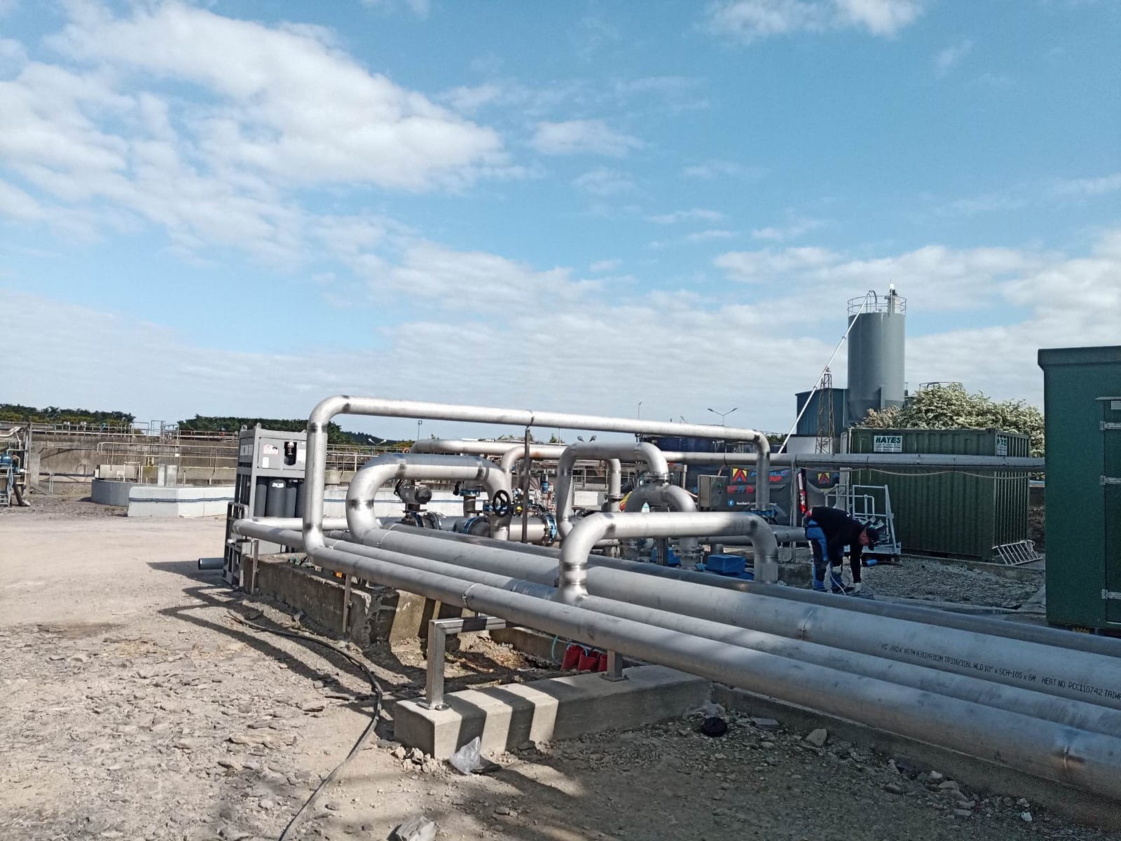 Instalacja przemysłowa z rur stalowych, widoczne zawory i armatura. W tle budynki przemysłowe i błękitne niebo z chmurami. Osoba w trakcie pracy przy instalacji.
