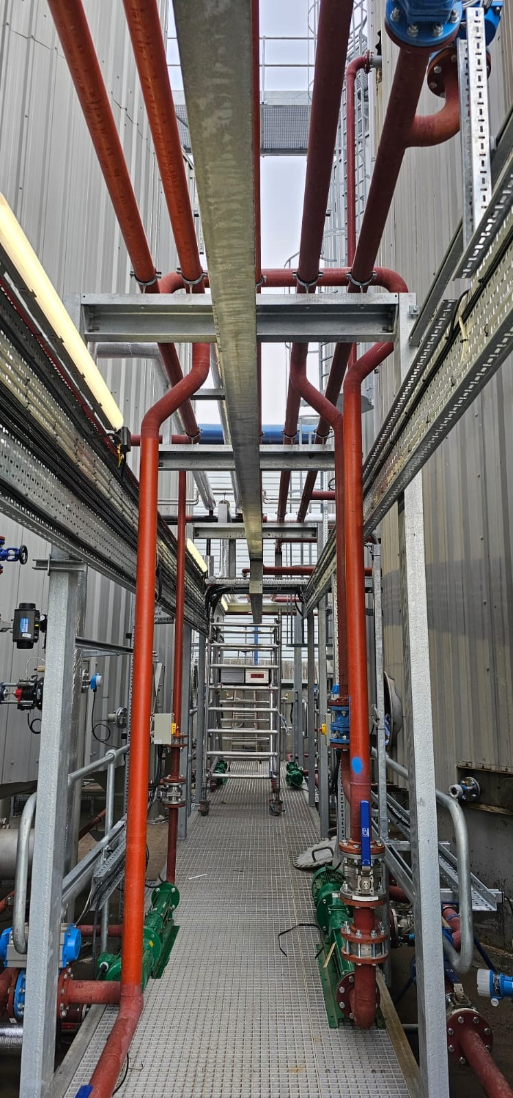 Instalacja przemysłowa z rurami w kolorze czerwonym i szarymi elementami konstrukcyjnymi. Widoczny fragment drabiny i metalowa podłoga kratowa.