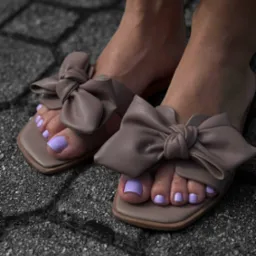 Stopa w sandałach z kokardą, paznokcie pomalowane na liliowo na tle szarej kostki brukowej. Delikatny, pastelowy odcień lakieru.